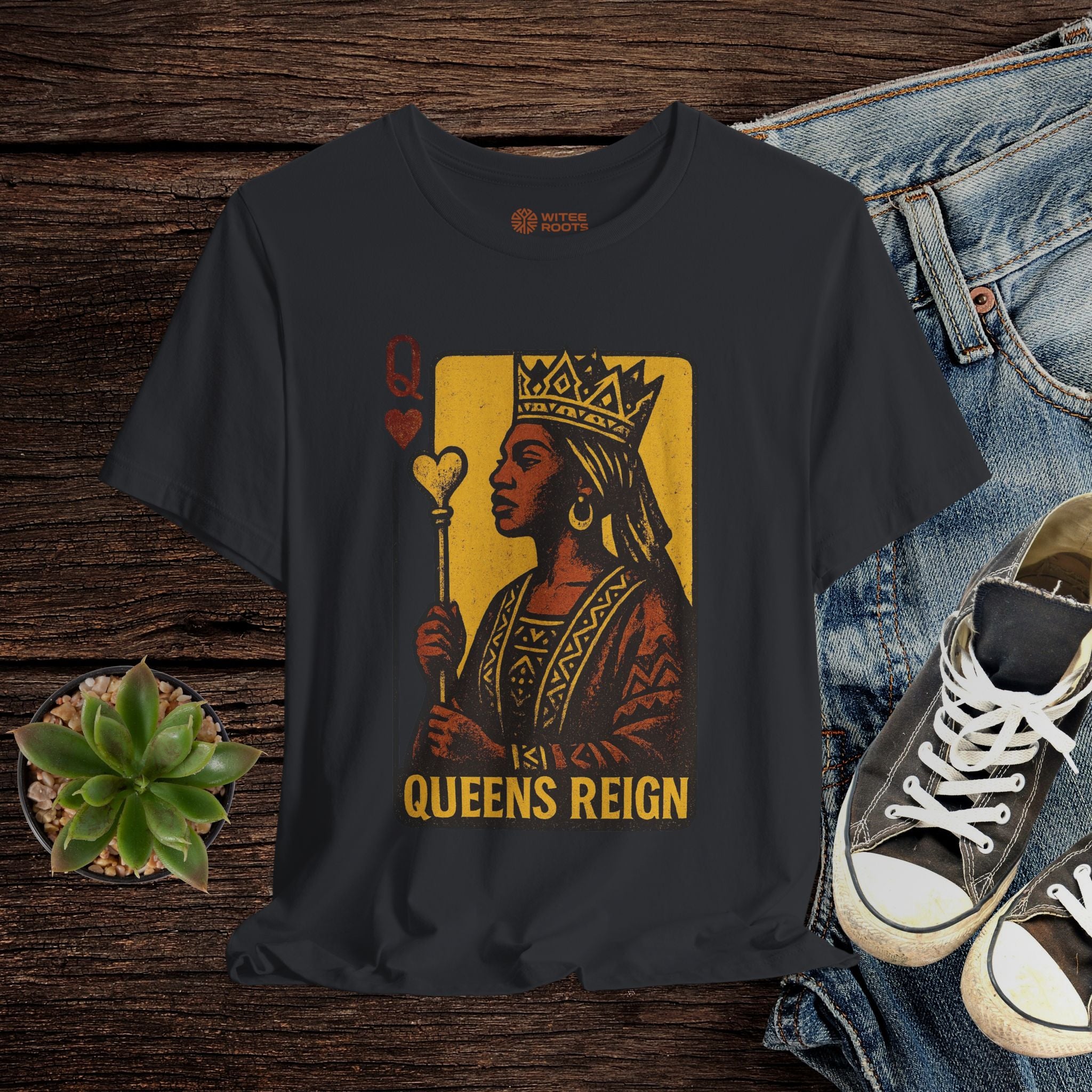 Black t-shirt with 'Queens Reign' design on a wooden surface with jeans and sneakers.