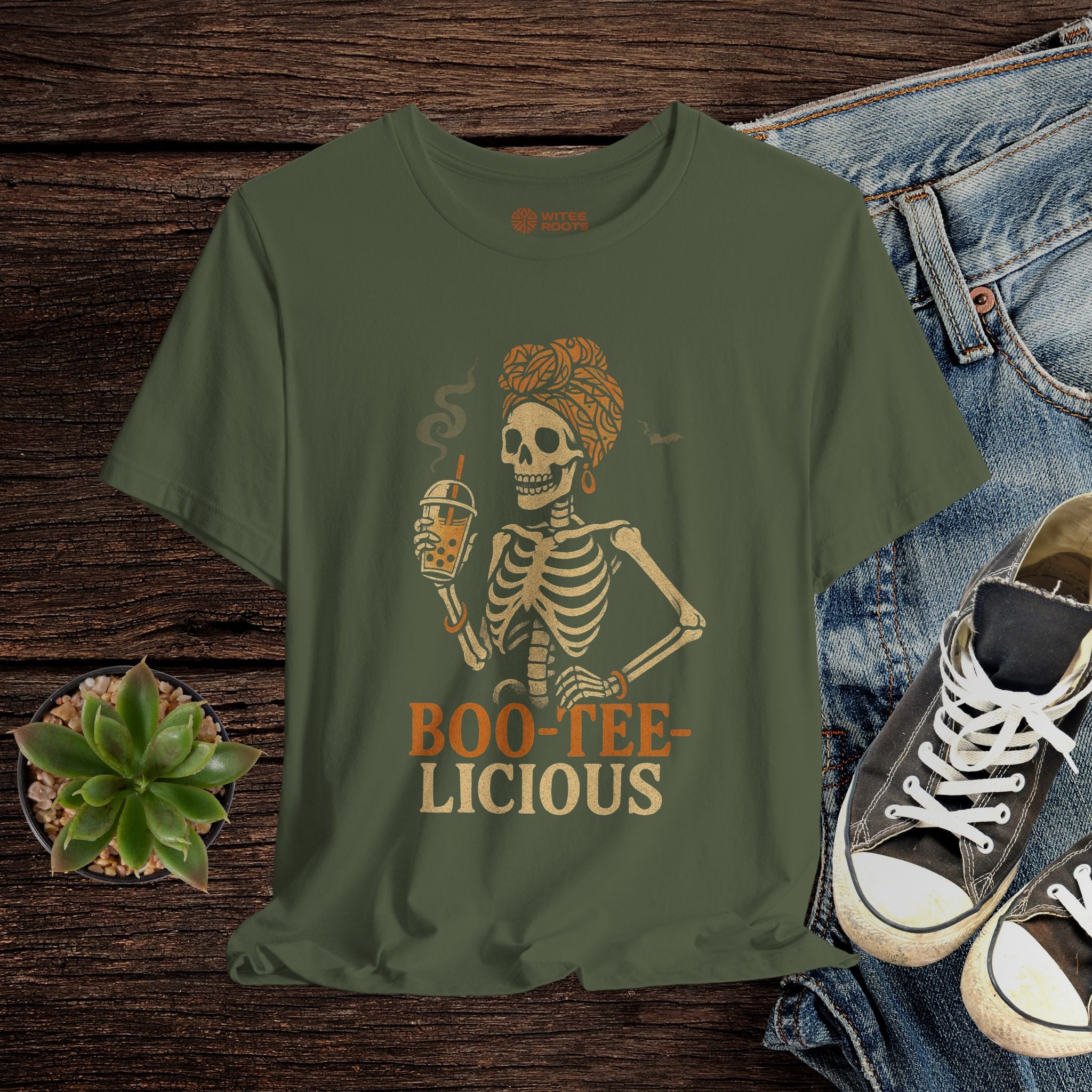 Green t-shirt with skeleton design and 'BOO-TEE-LICIOUS' text on a wooden surface with jeans and sneakers.