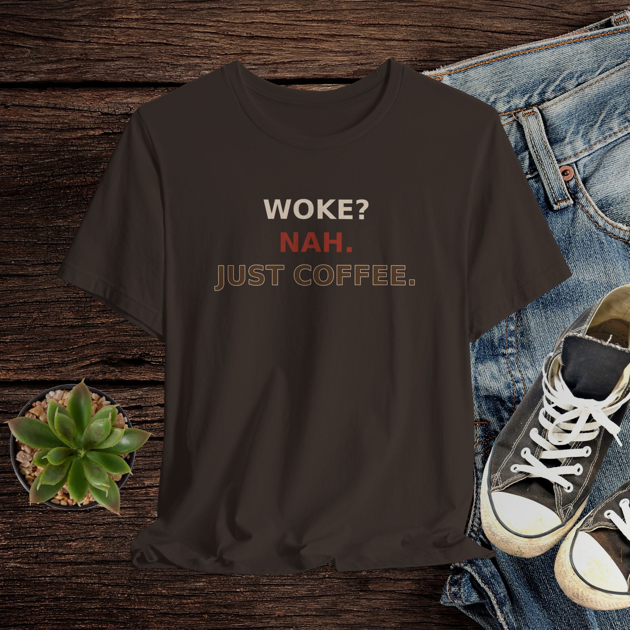 Brown t-shirt with 'WOKE? NAH. JUST COFFEE.' text on a wooden surface with jeans and sneakers.