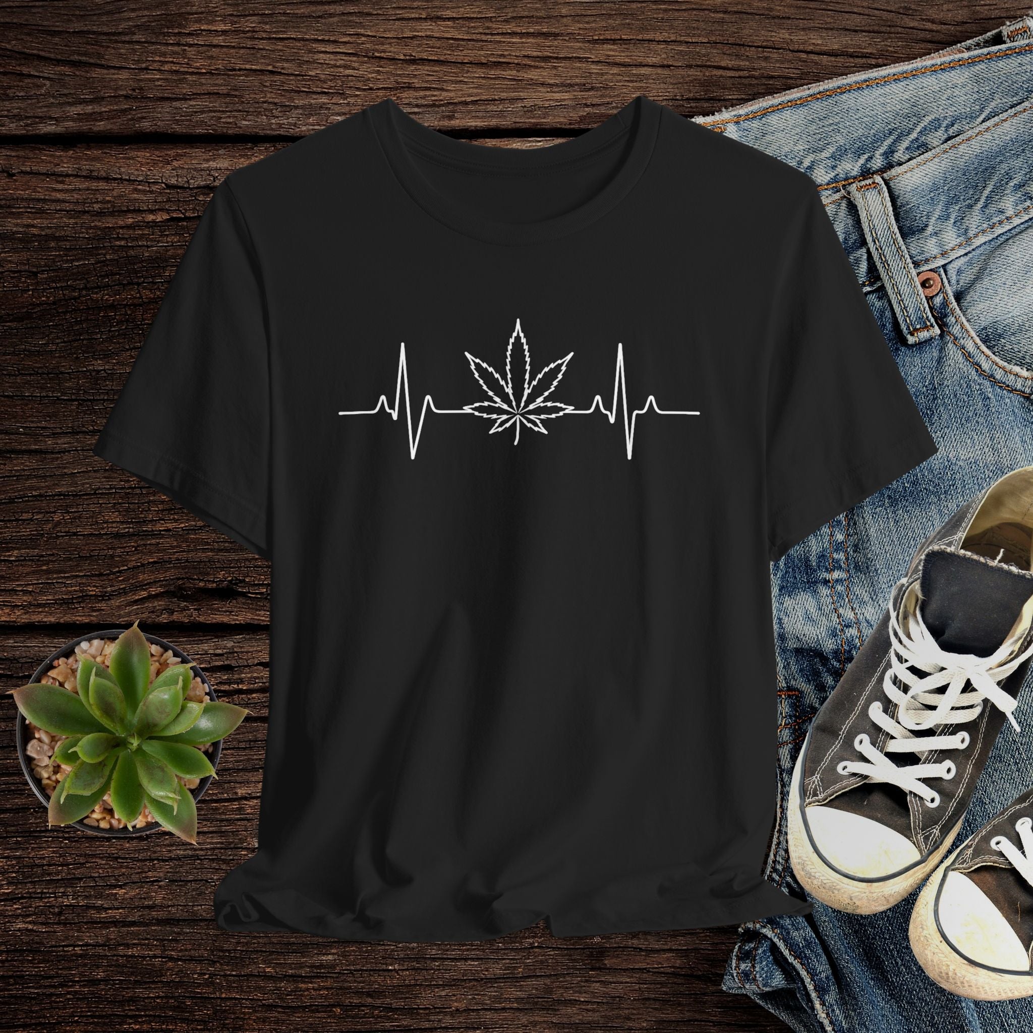 Black t-shirt with a cannabis leaf and heartbeat line design on a wooden surface with jeans and sneakers.