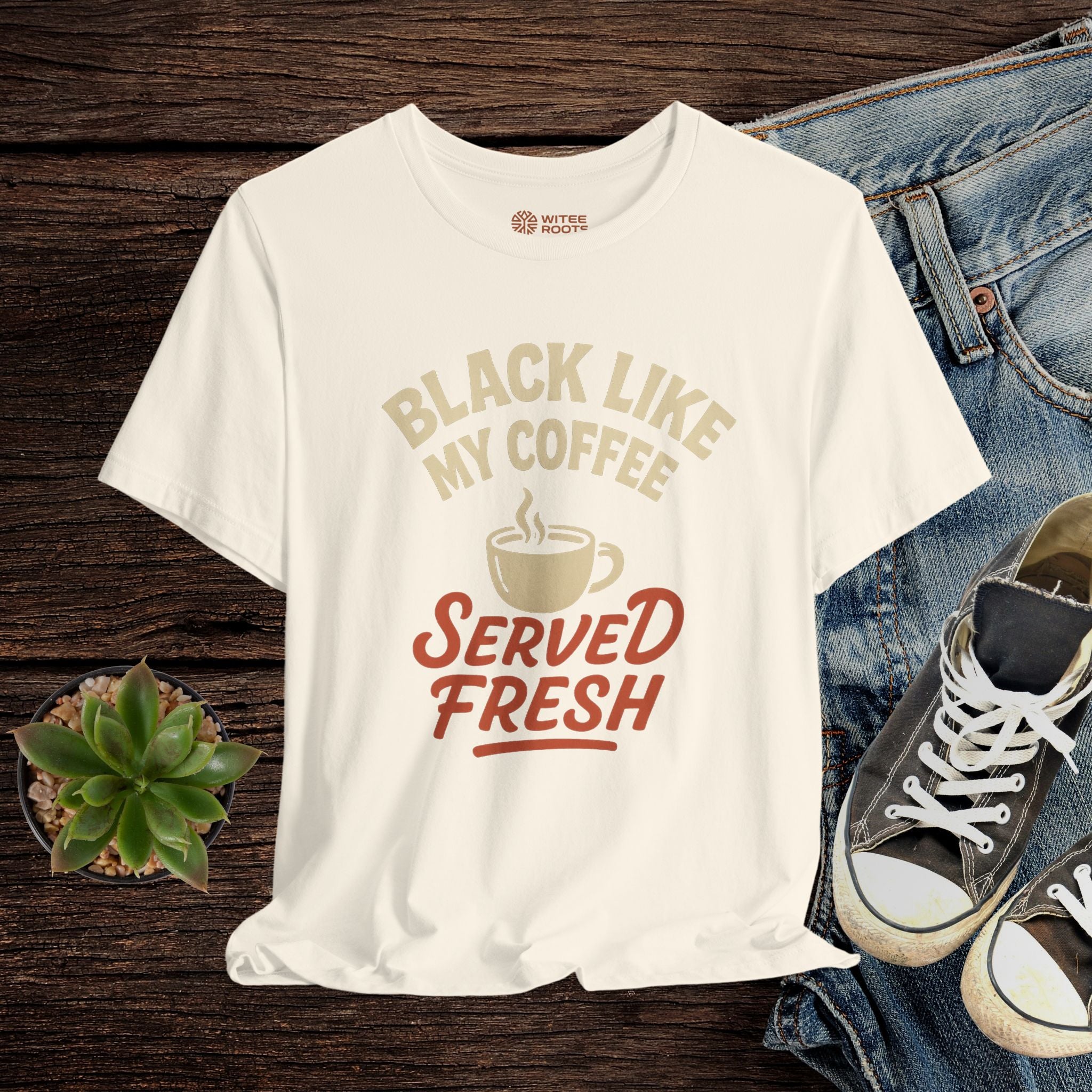 White t-shirt with coffee-themed text and graphics on a wooden surface with jeans and sneakers.