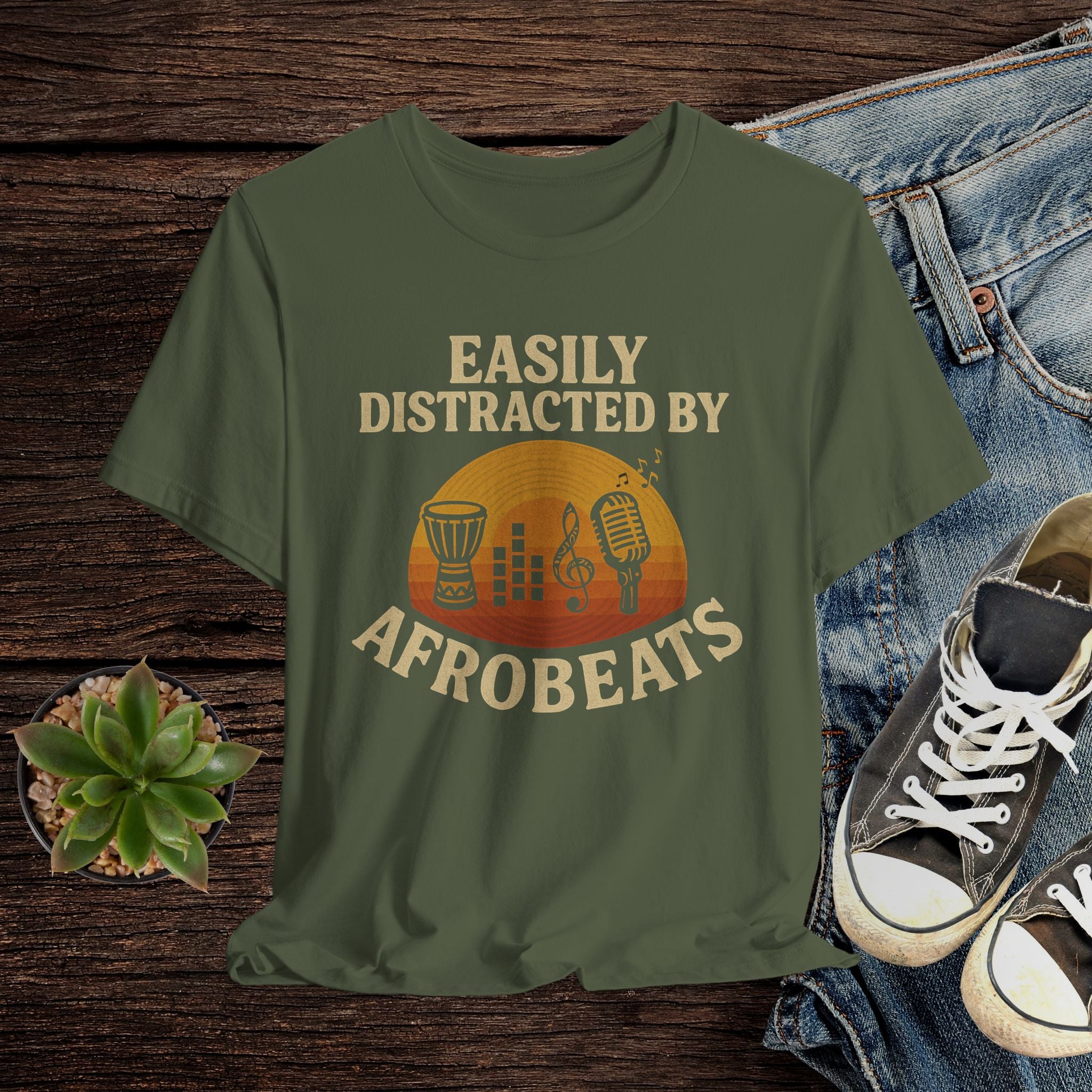 Green t-shirt with 'Easily Distracted by Afrobeats' design on a wooden surface with jeans and sneakers.