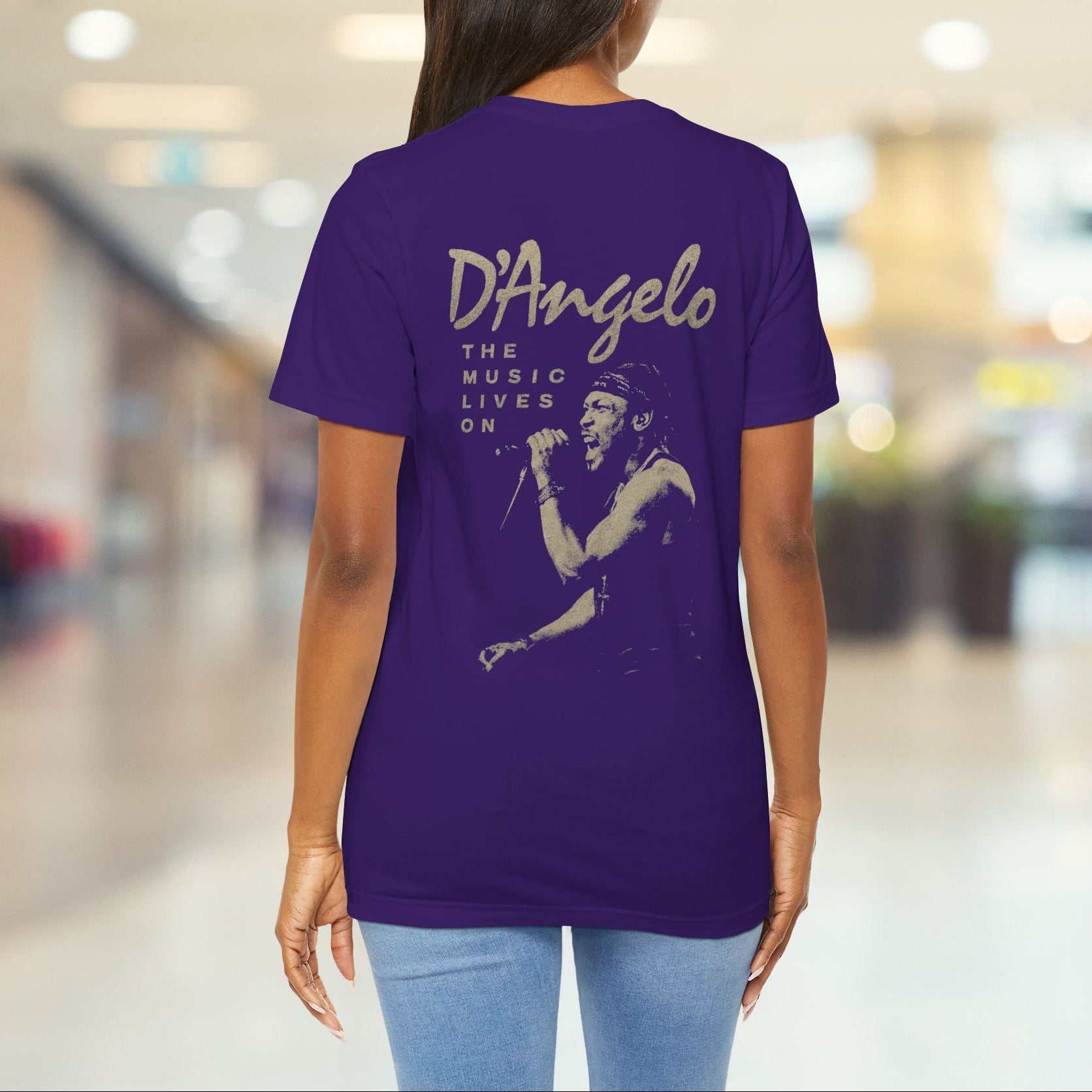 Person wearing a purple t-shirt with 'D'Angelo The Music Lives On' design in a blurred indoor setting