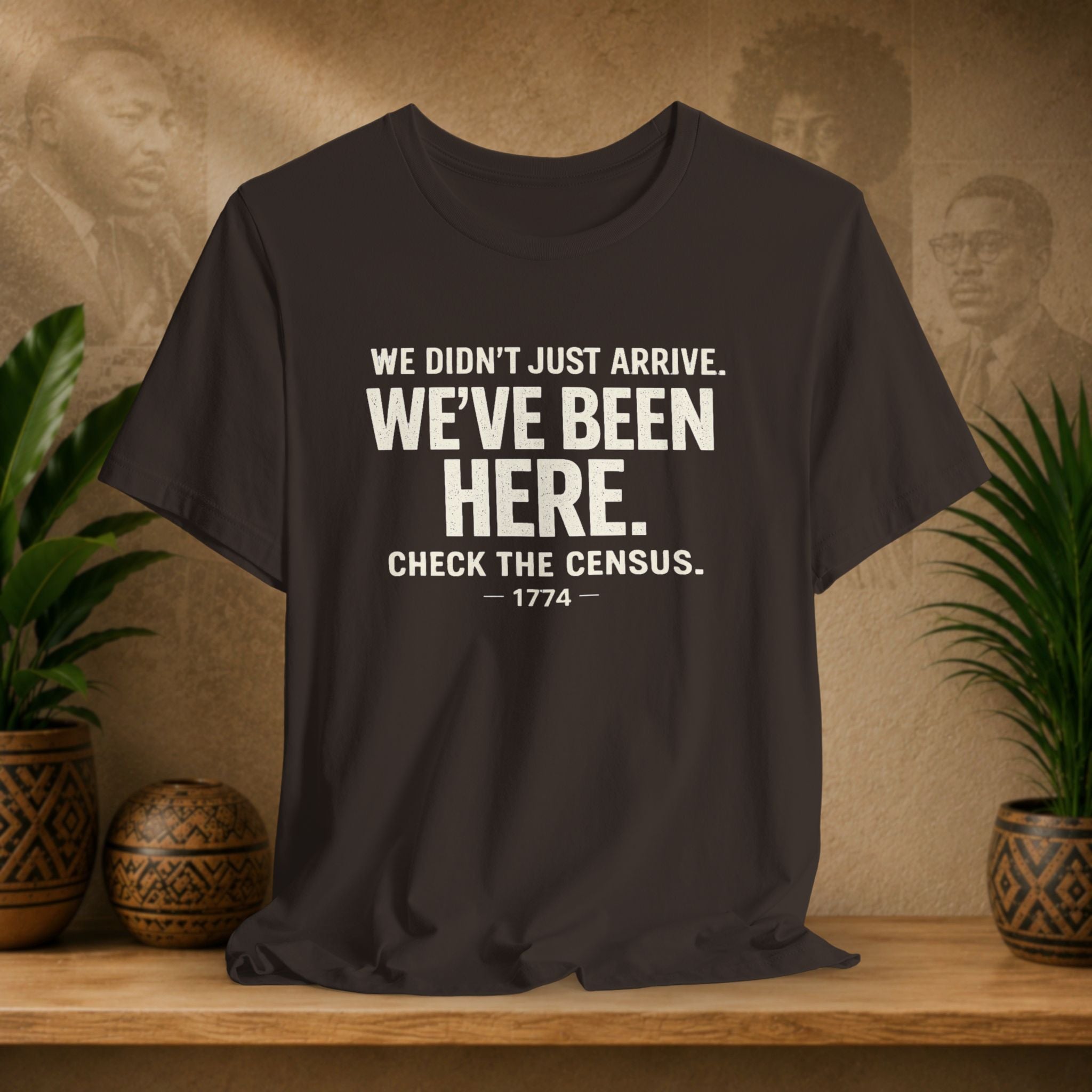 Brown t-shirt with white text on a wooden surface with plants and a textured wall background