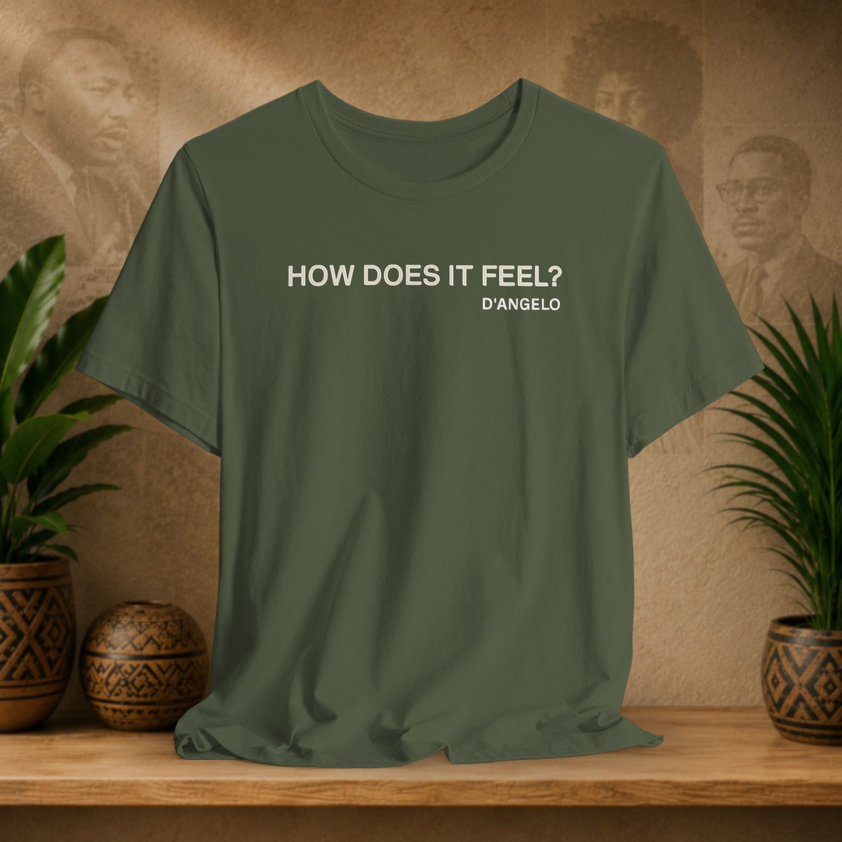 Green t-shirt with 'HOW DOES IT FEEL? D'ANGELO' text on a wooden surface with decorative plants and background.