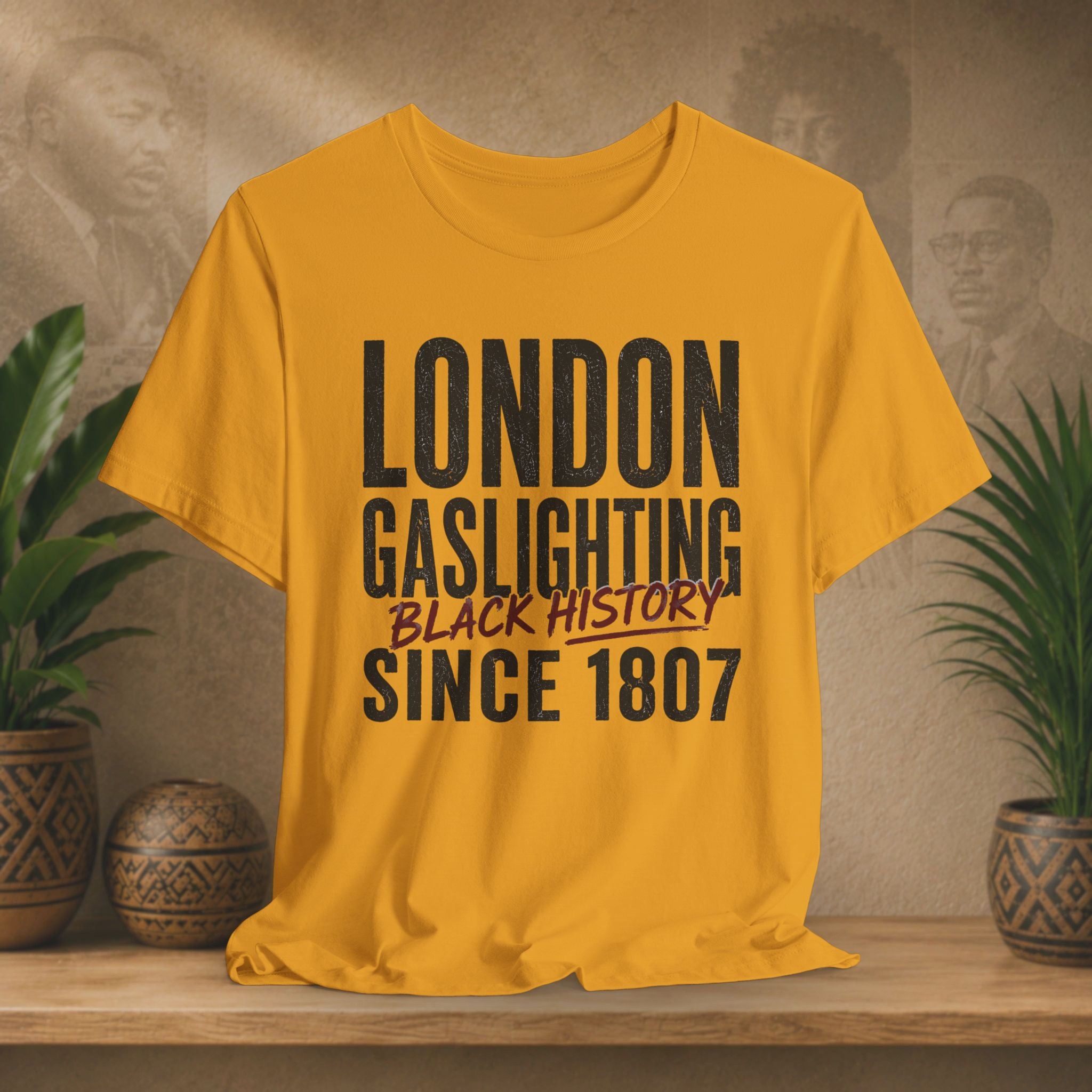 Mustard t-shirt with 'London Gaslighting Black History Since 1807' text on a wooden surface with plants and a blurred background.