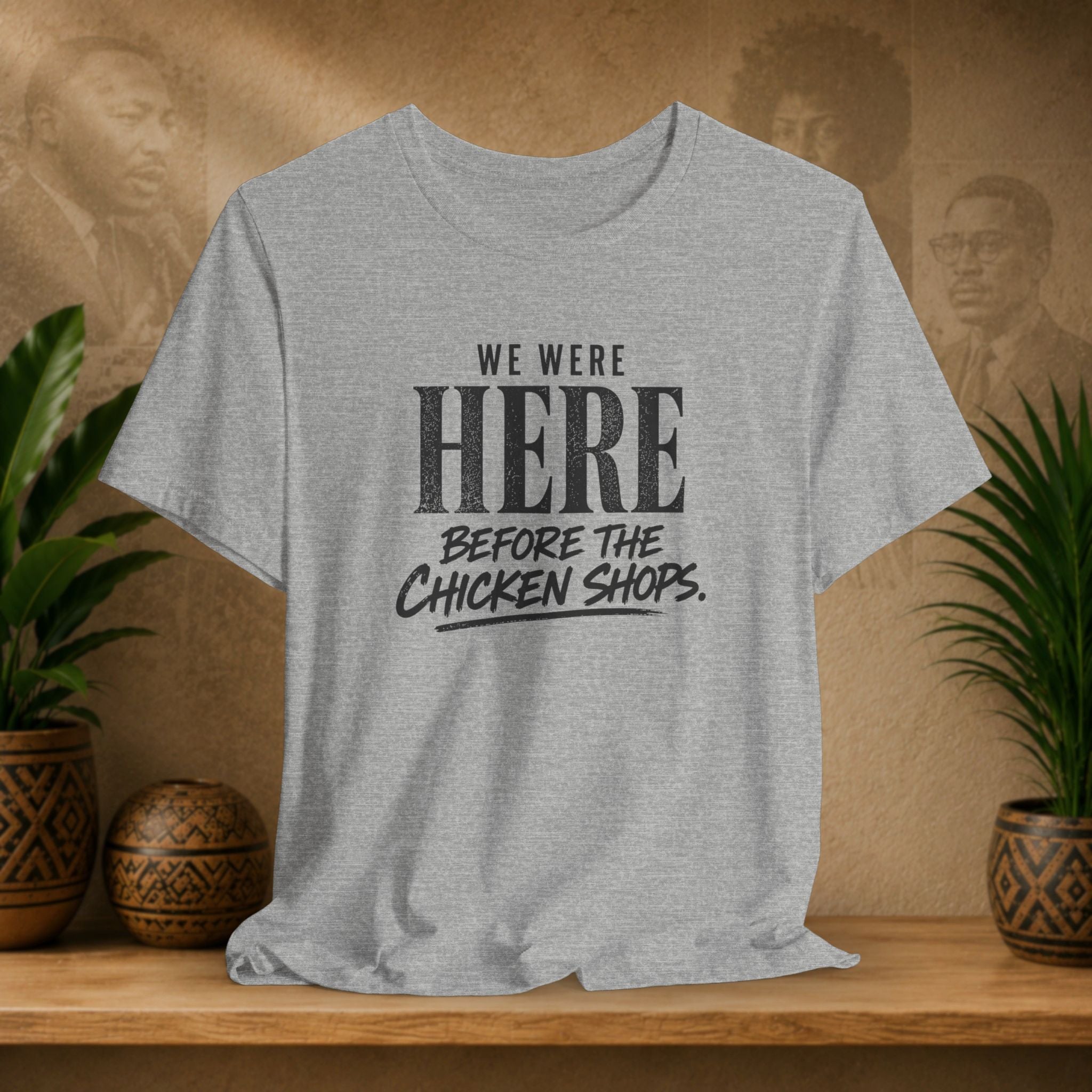 Gray t-shirt with text 'We were here before the chicken shops' on a wooden surface with plants and a textured wall background.