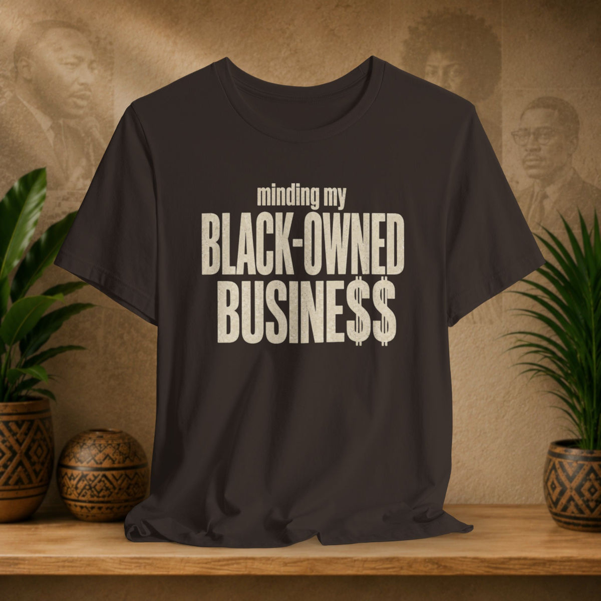 A brown t-shirt with the text 'minding my BLACK-OWNED BUSINESS$$' printed in distressed cream lettering, displayed on a wooden shelf with potted plants against a textured beige background.