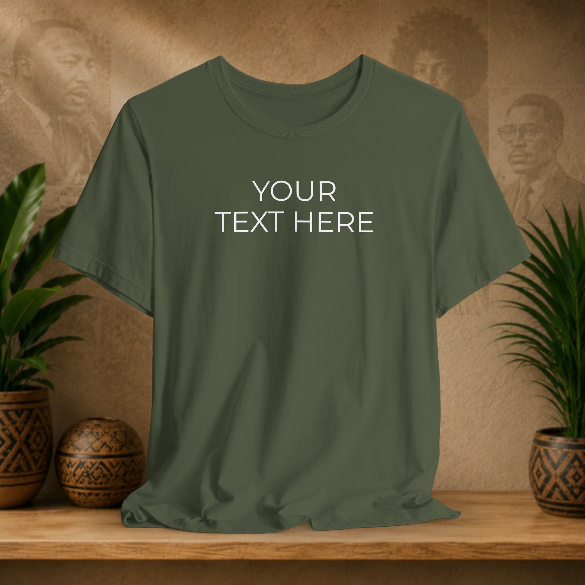 An olive green short-sleeve t-shirt with 'YOUR TEXT HERE' printed in white on the chest, displayed on a wooden shelf with potted plants and a textured background.