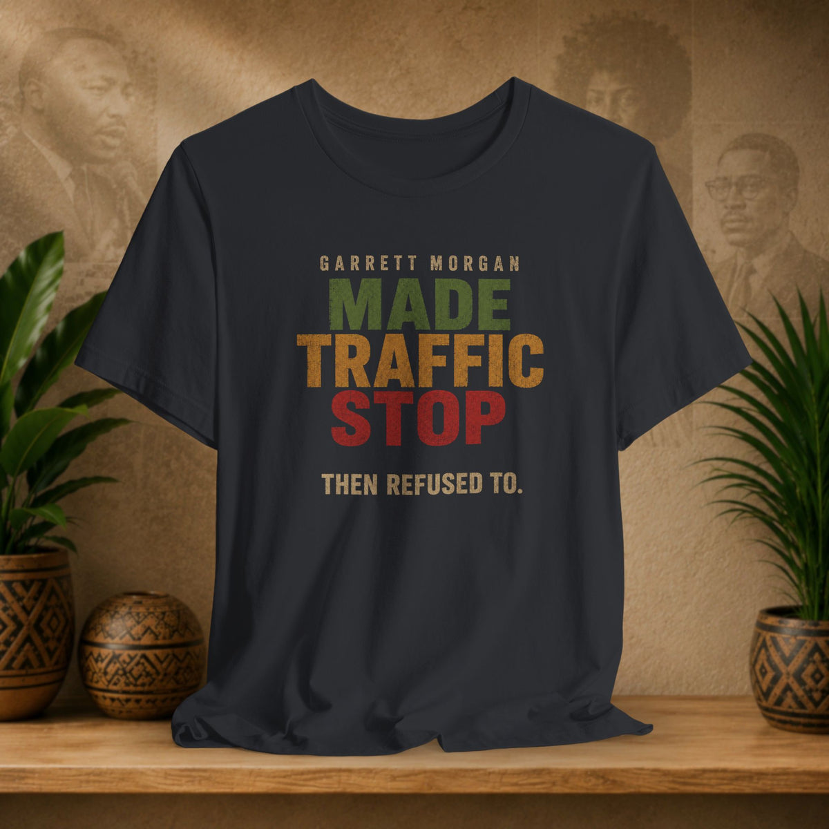 A black graphic t-shirt displayed on a wooden shelf. The shirt features a retro-style text graphic that reads 'GARRETT MORGAN MADE TRAFFIC STOP THEN REFUSED TO.