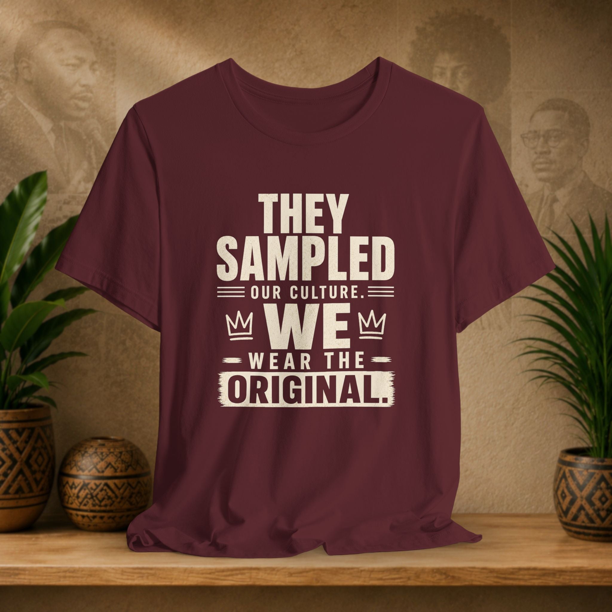 Maroon t-shirt with white text on a wooden surface with plants and a wall with portraits in the background