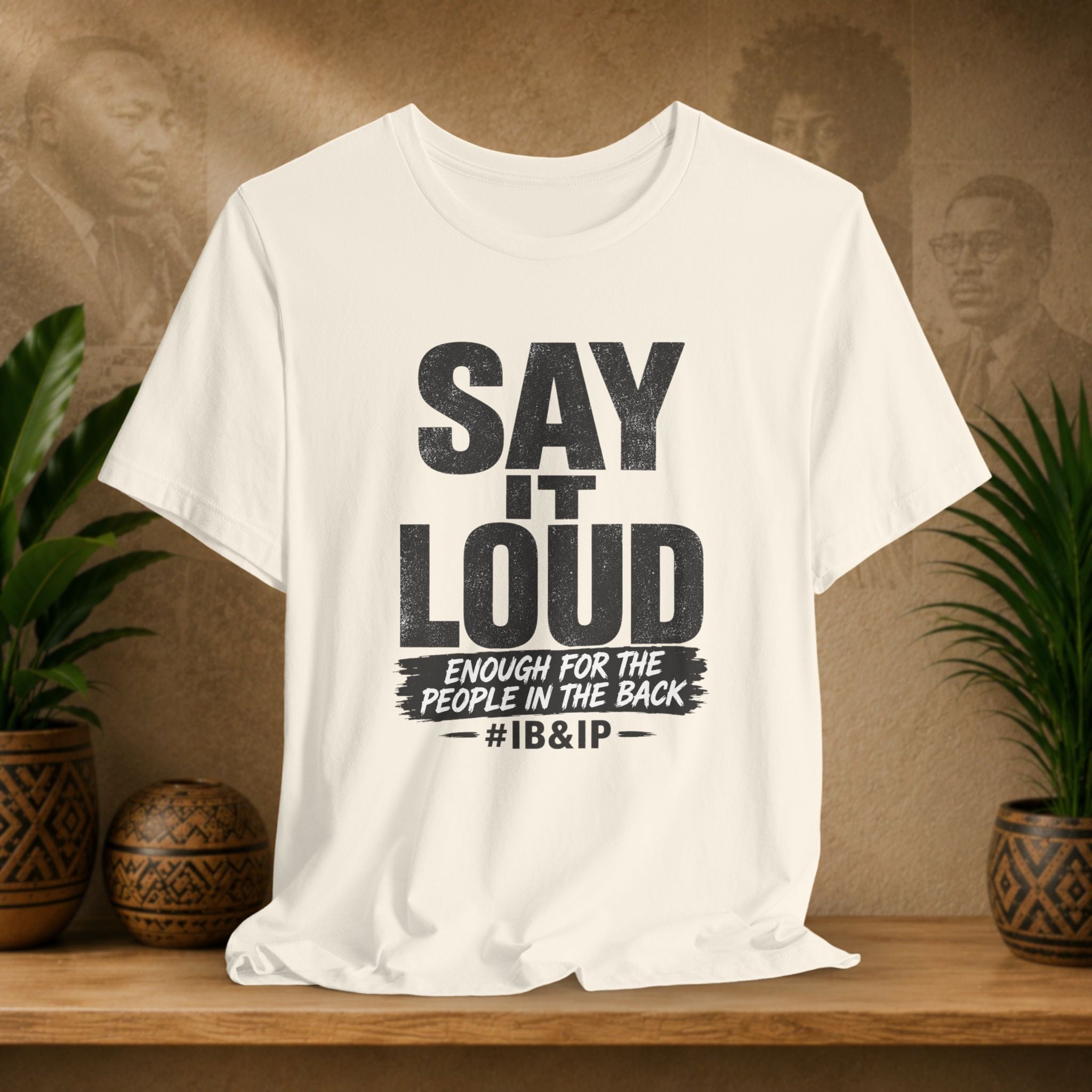 White t-shirt with text 'Say it Loud Enough for the People in the Back #IB&IP' on a wooden surface with plants and a textured wall background.