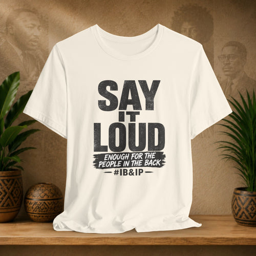 White t-shirt with text 'Say it Loud Enough for the People in the Back #IB&IP' on a wooden surface with plants and a textured wall background.