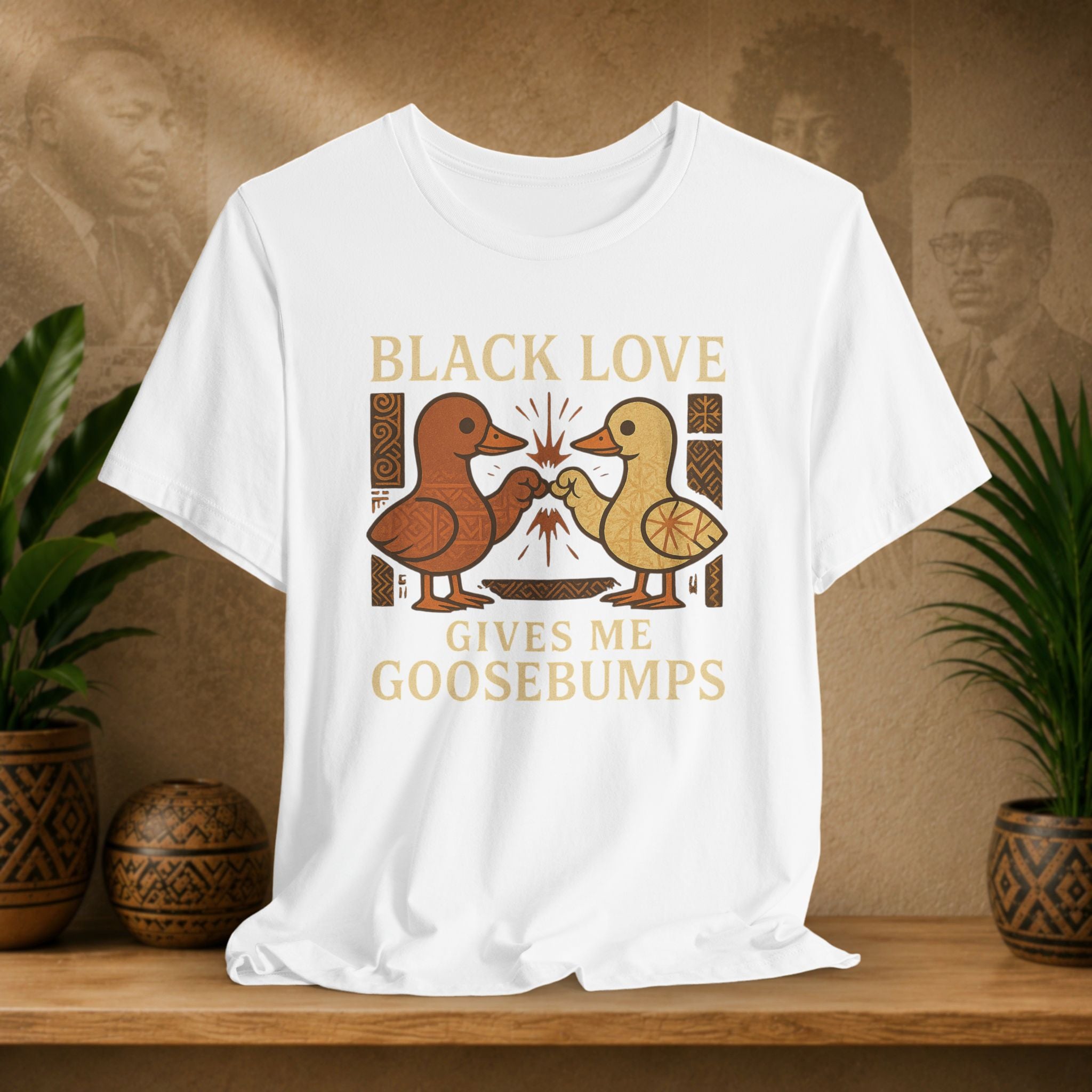 White t-shirt with 'Black Love Gives Me Goosebumps' graphic on a wooden surface with plants and a textured wall background.