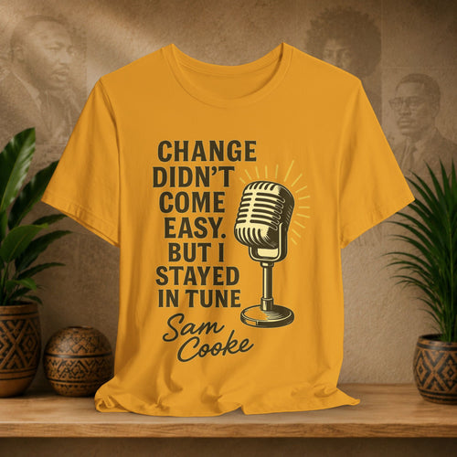 Mustard t-shirt with microphone graphic and text on a wooden surface with plants and wall art.