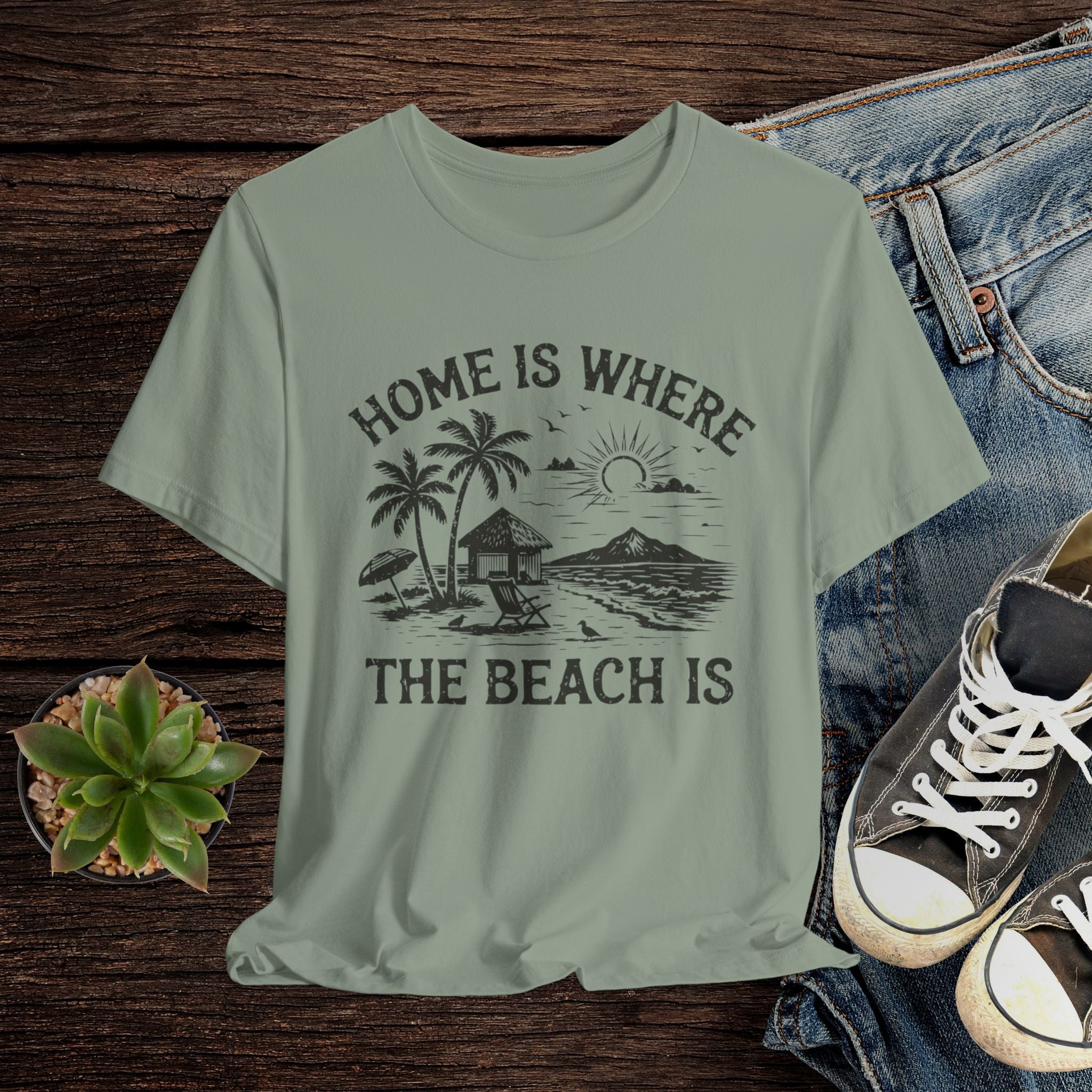 Sage green t-shirt with beach graphic and text on a wooden surface with jeans and sneakers.