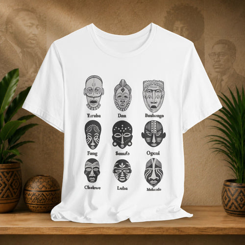 White t-shirt with black tribal mask designs and text on a wooden surface with plants and decorative items.