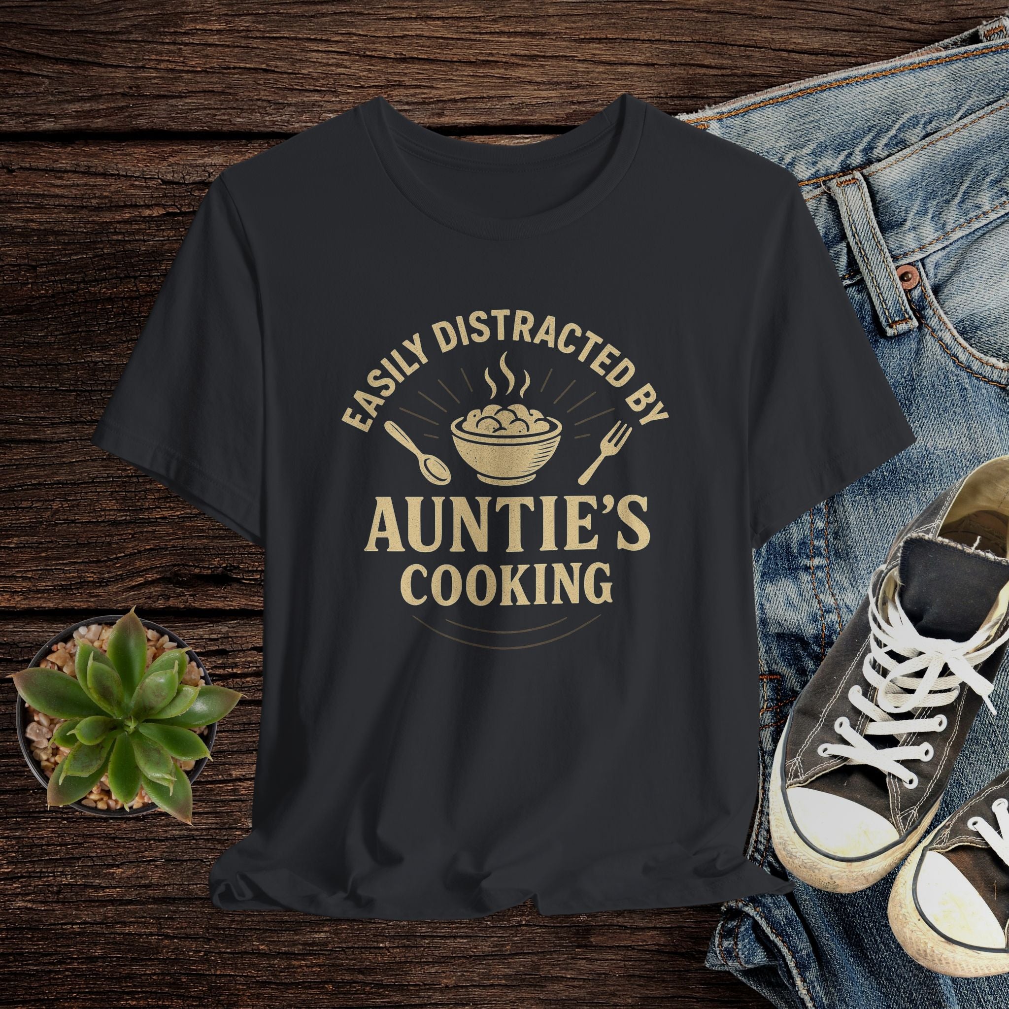 Black t-shirt with 'Easily Distracted by Auntie's Cooking' text on a wooden surface with jeans and sneakers.
