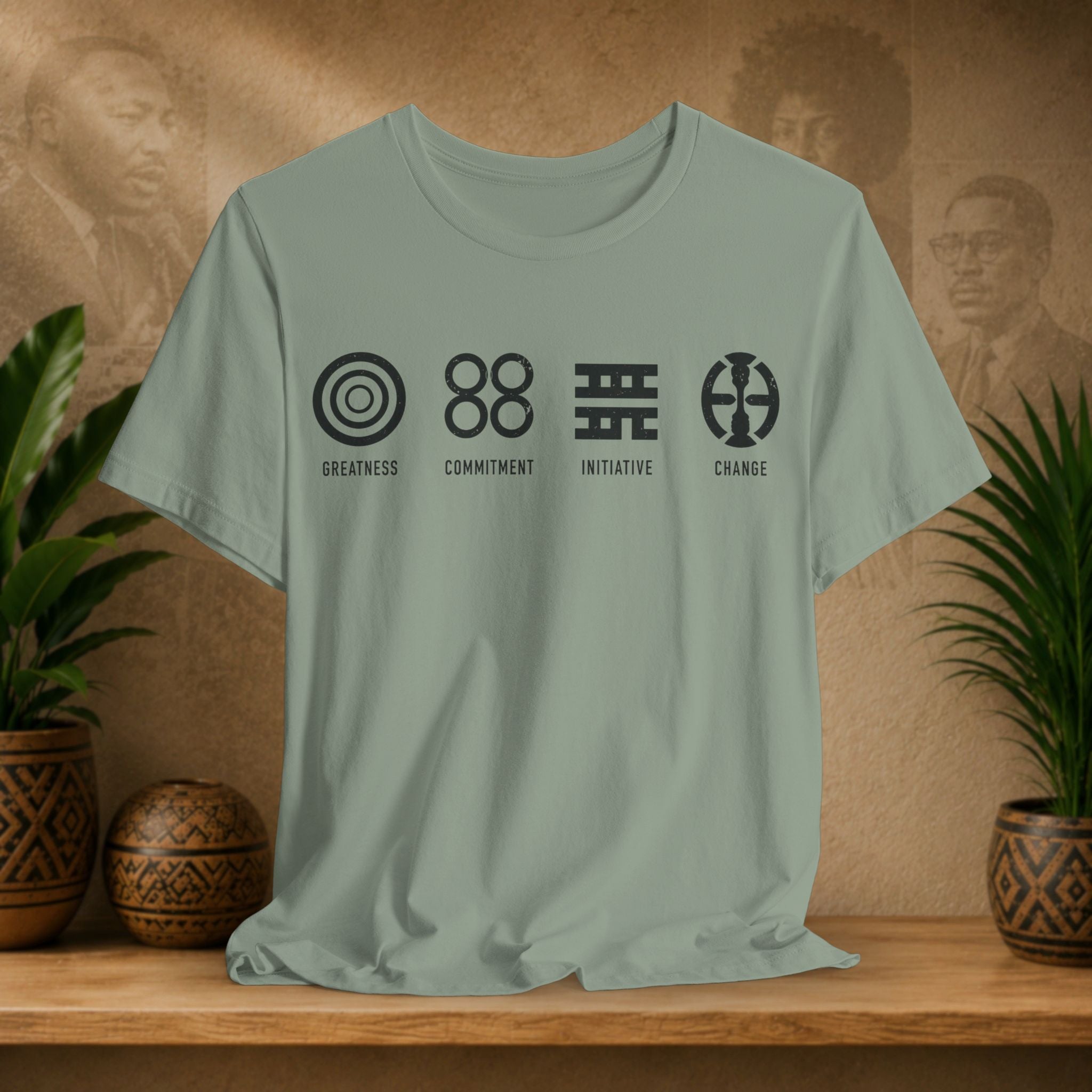 Sage green t-shirt with black symbols and text on a wooden surface with plants and a textured wall background