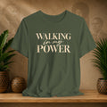 An olive green short-sleeve t-shirt with the text 'WALKING in my POWER' printed in beige on the front, displayed on a wooden shelf with potted plants and a textured background.