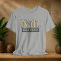 Gray t-shirt with 'Nah. Rosa Parks' text on a wooden shelf with plants and a blurred background.