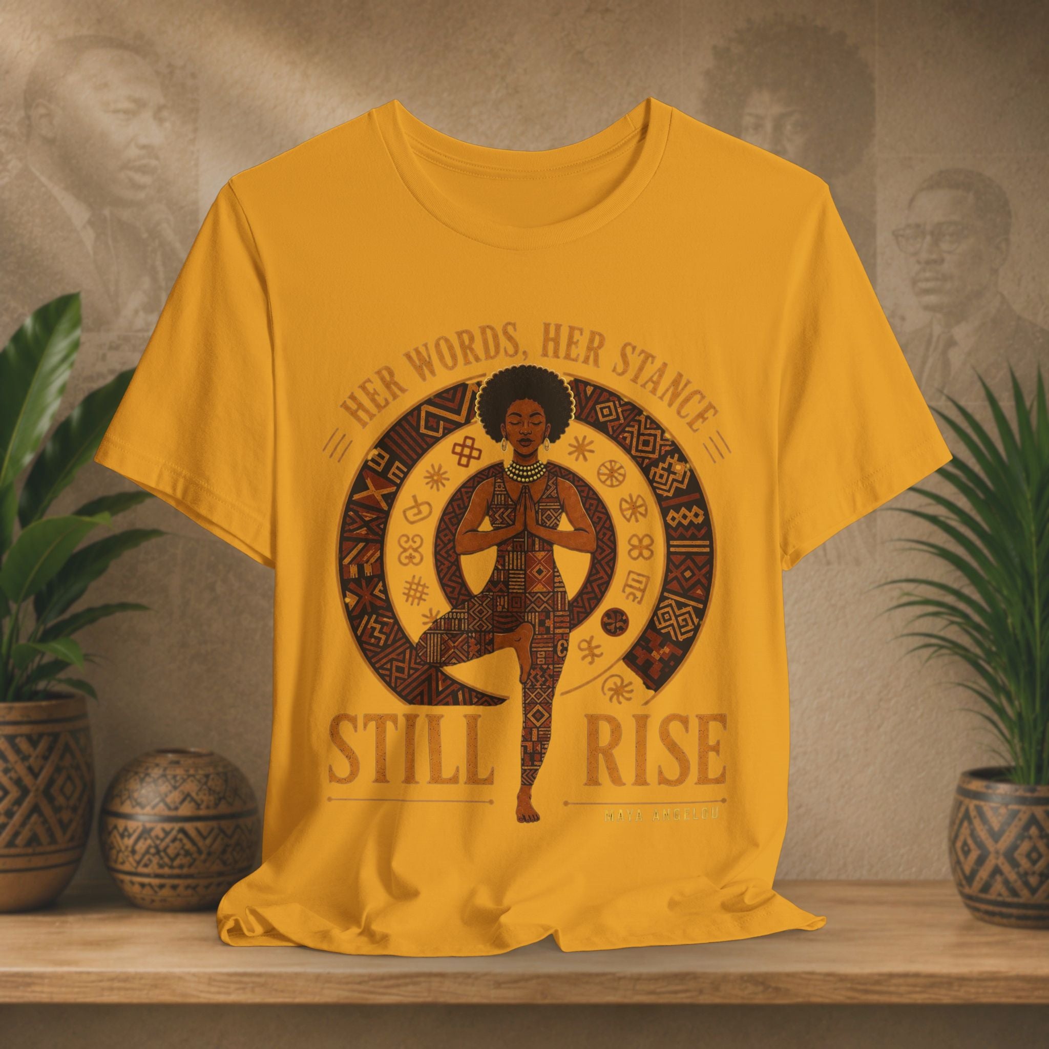 A mustard yellow graphic t-shirt displayed on a wooden shelf, flanked by potted plants. The shirt features a brown and black graphic of a woman in a yoga pose inside a circular pattern, with the text 'HER WORDS, HER STANCE STILL RISE' printed around her.
