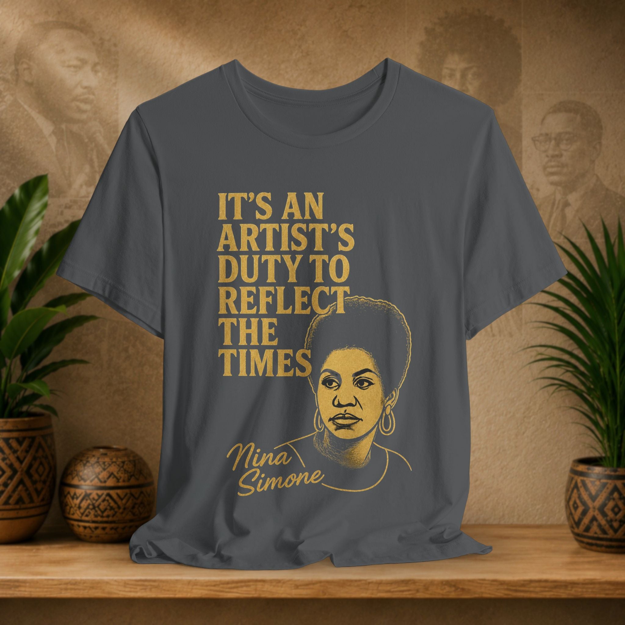 Gray t-shirt with gold text and illustration of a woman, placed on a wooden surface with a decorative background.