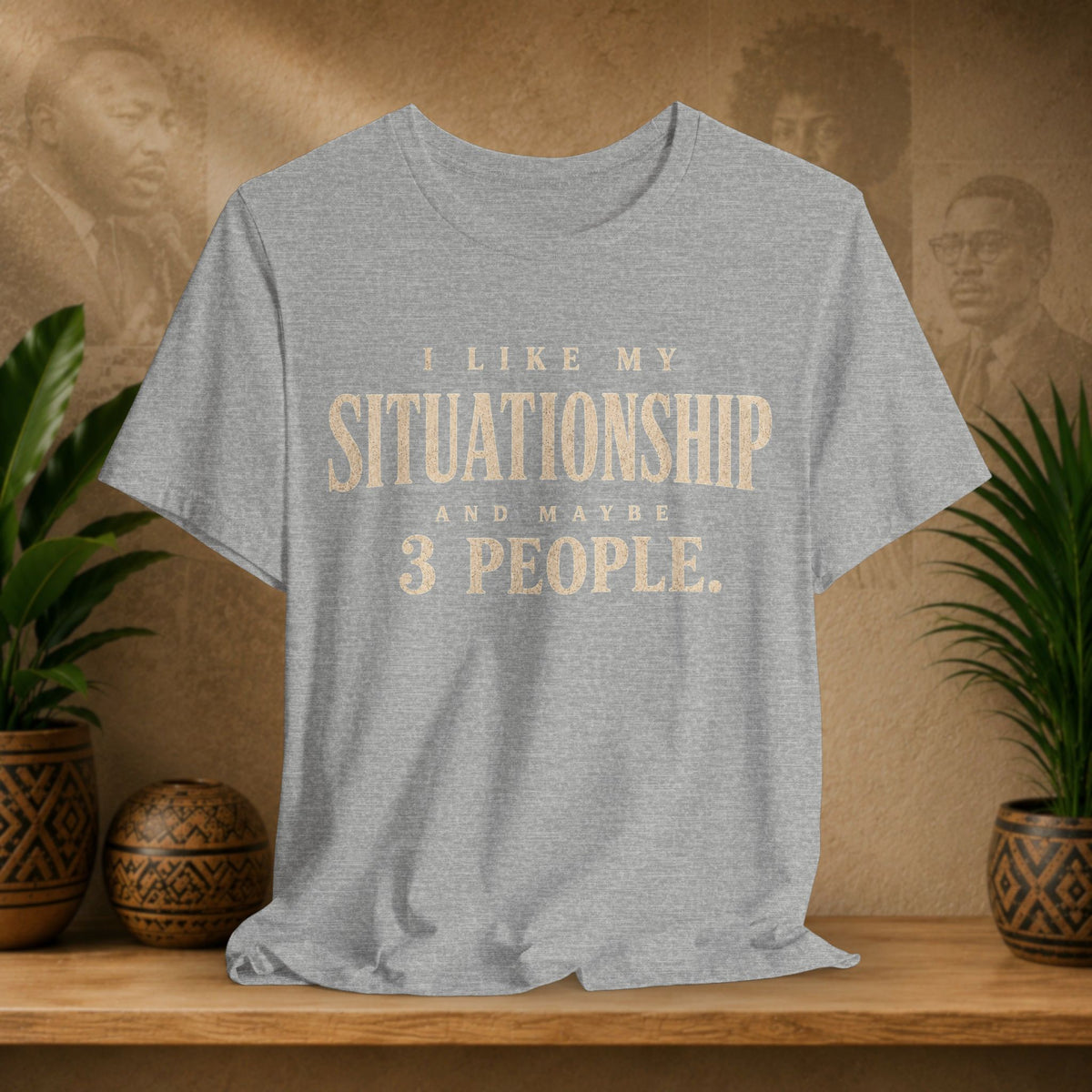 A heather gray short-sleeve t-shirt with a distressed tan text graphic reading 'I LIKE MY SITUATIONSHIP AND MAYBE 3 PEOPLE.' displayed on a wooden shelf with potted plants and a textured background.