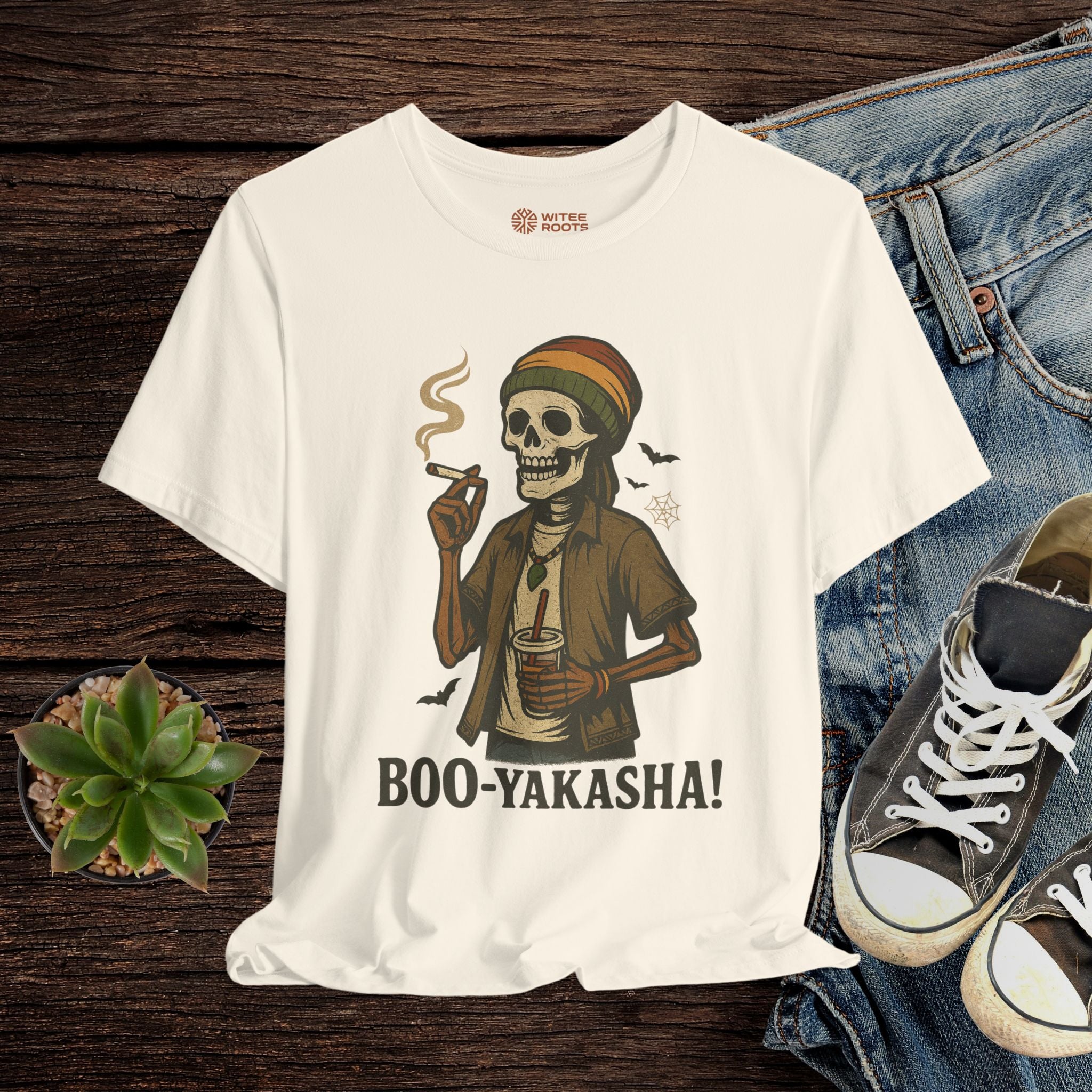 Beige t-shirt with a skeleton wearing a hat and smoking a cigarette, displayed with jeans and sneakers on a wooden surface.