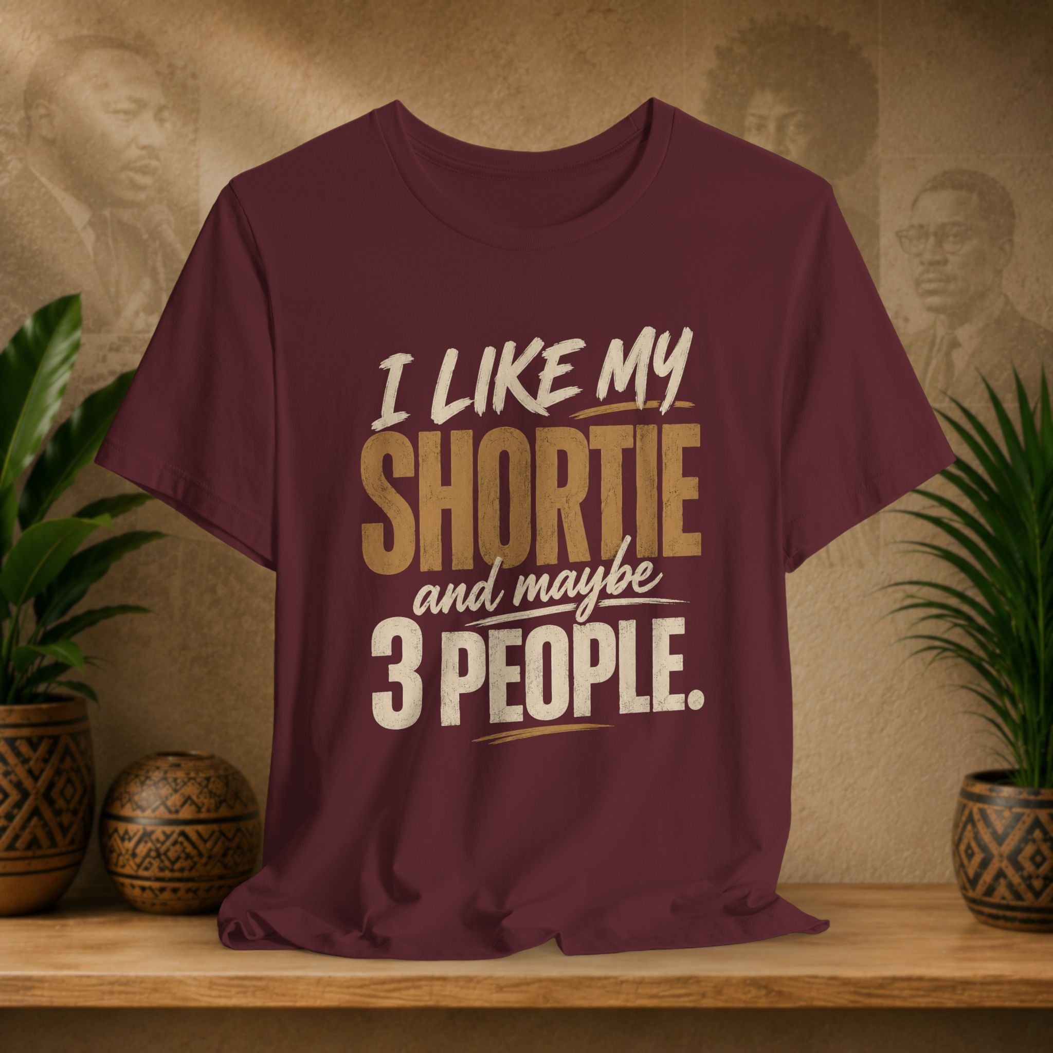 A maroon graphic t-shirt displayed on a wooden shelf. The shirt features a humorous slogan printed in white and tan lettering: 'I LIKE MY SHORTIE and maybe 3 PEOPLE.