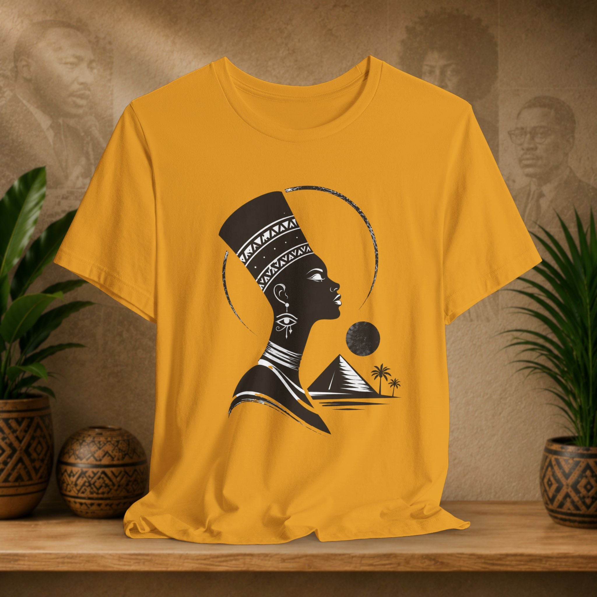 Yellow t-shirt with a black silhouette design of a woman with a headdress, pyramid, and palm trees on a wooden surface with plants and decorative items.