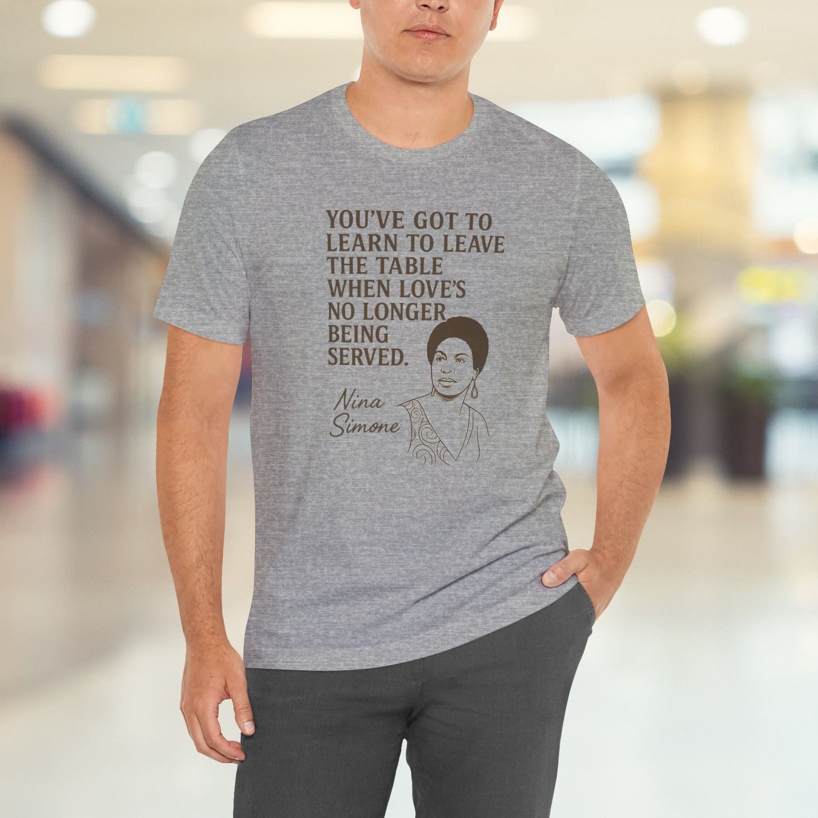 Person wearing a gray t-shirt with a quote and a portrait, standing in a blurred indoor setting.