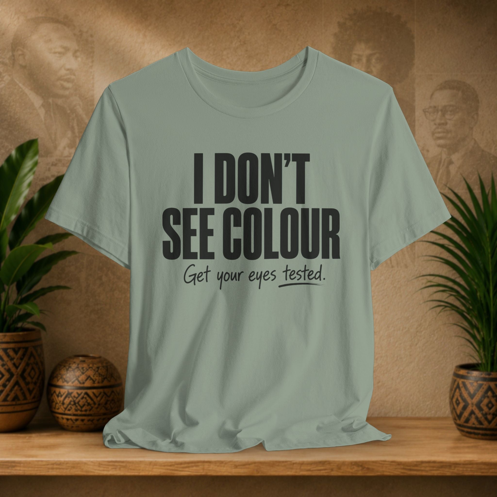 Sage green t-shirt with text 'I DON'T SEE COLOUR Get your eyes tested.' on a wooden surface with plants and a blurred background.