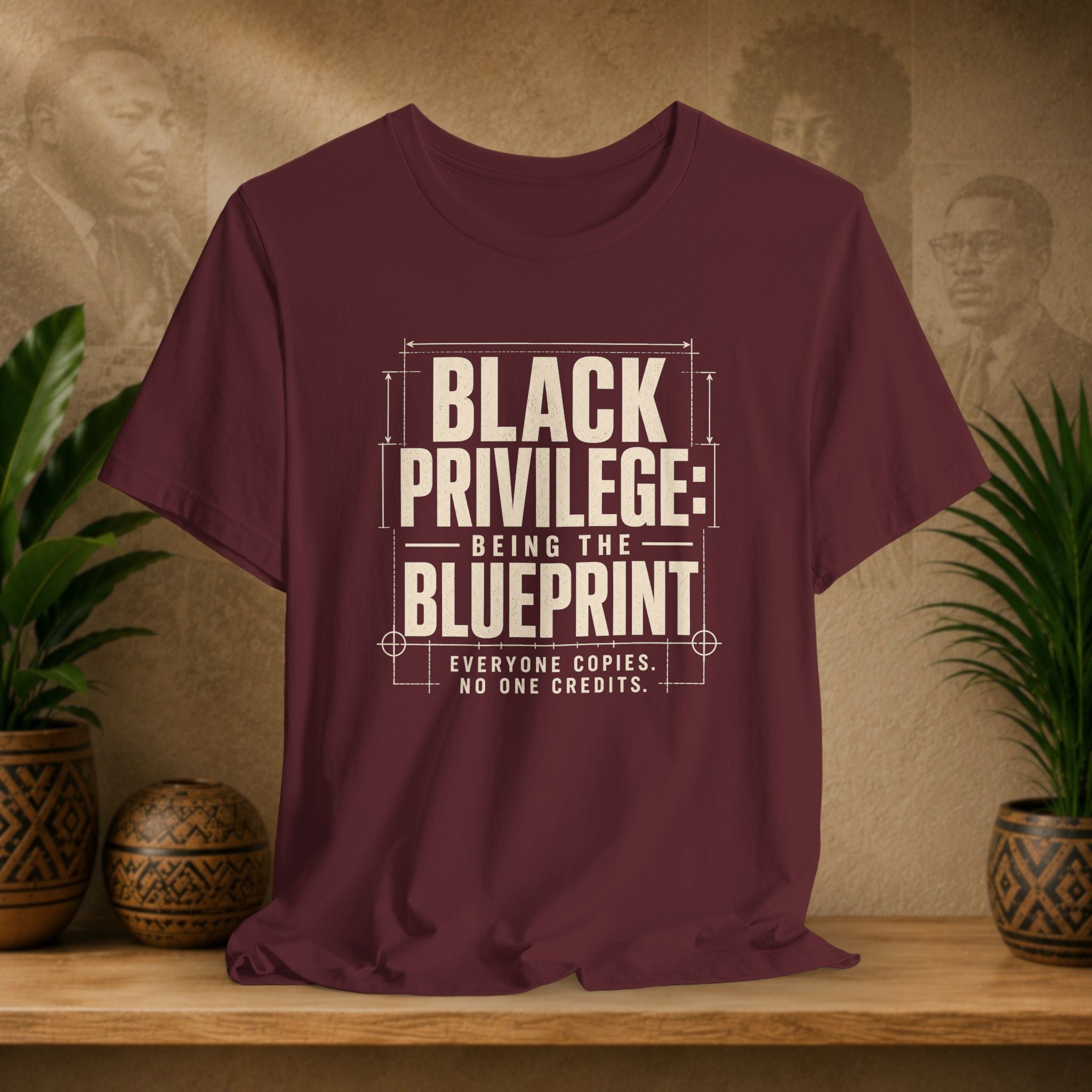 Maroon t-shirt with white text on a wooden surface with plants and a textured wall background