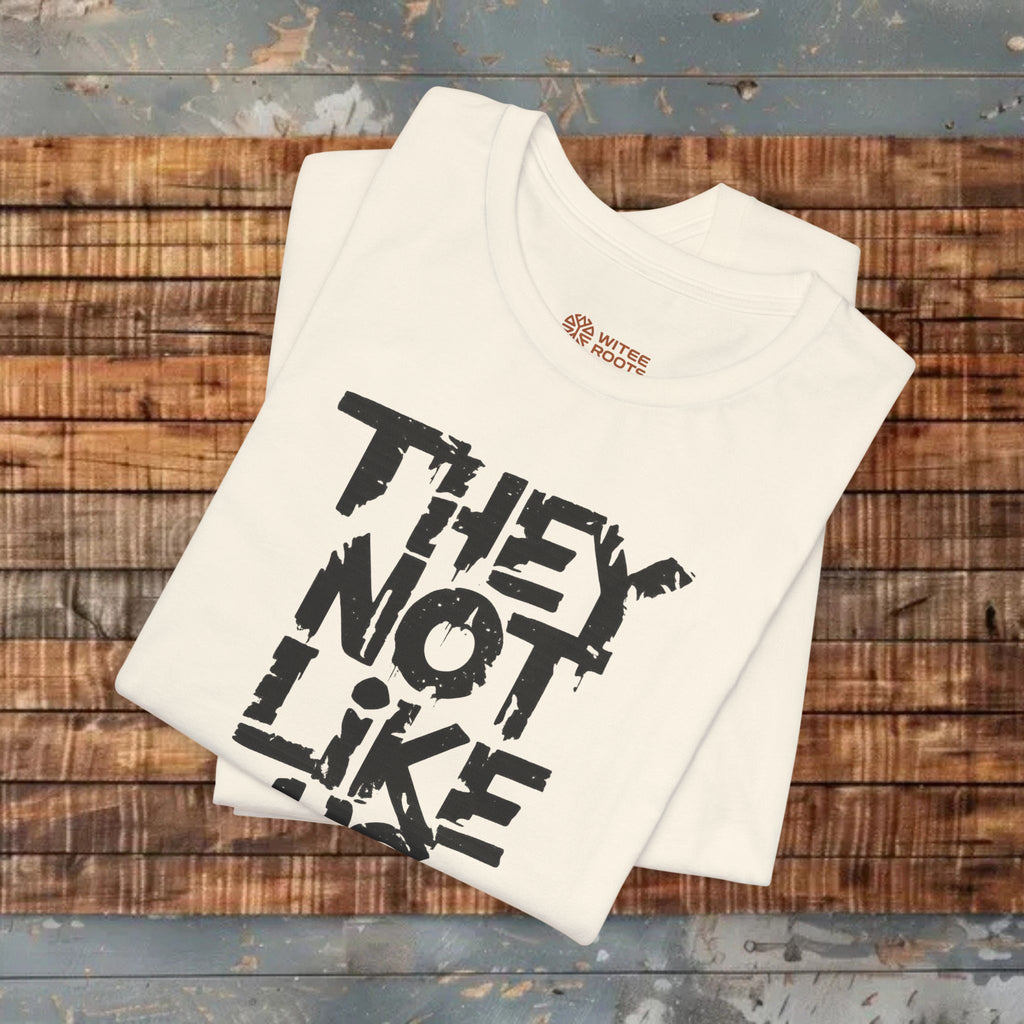 Beige t-shirt with black text 'THEY NOT LIKE' on a wooden surface