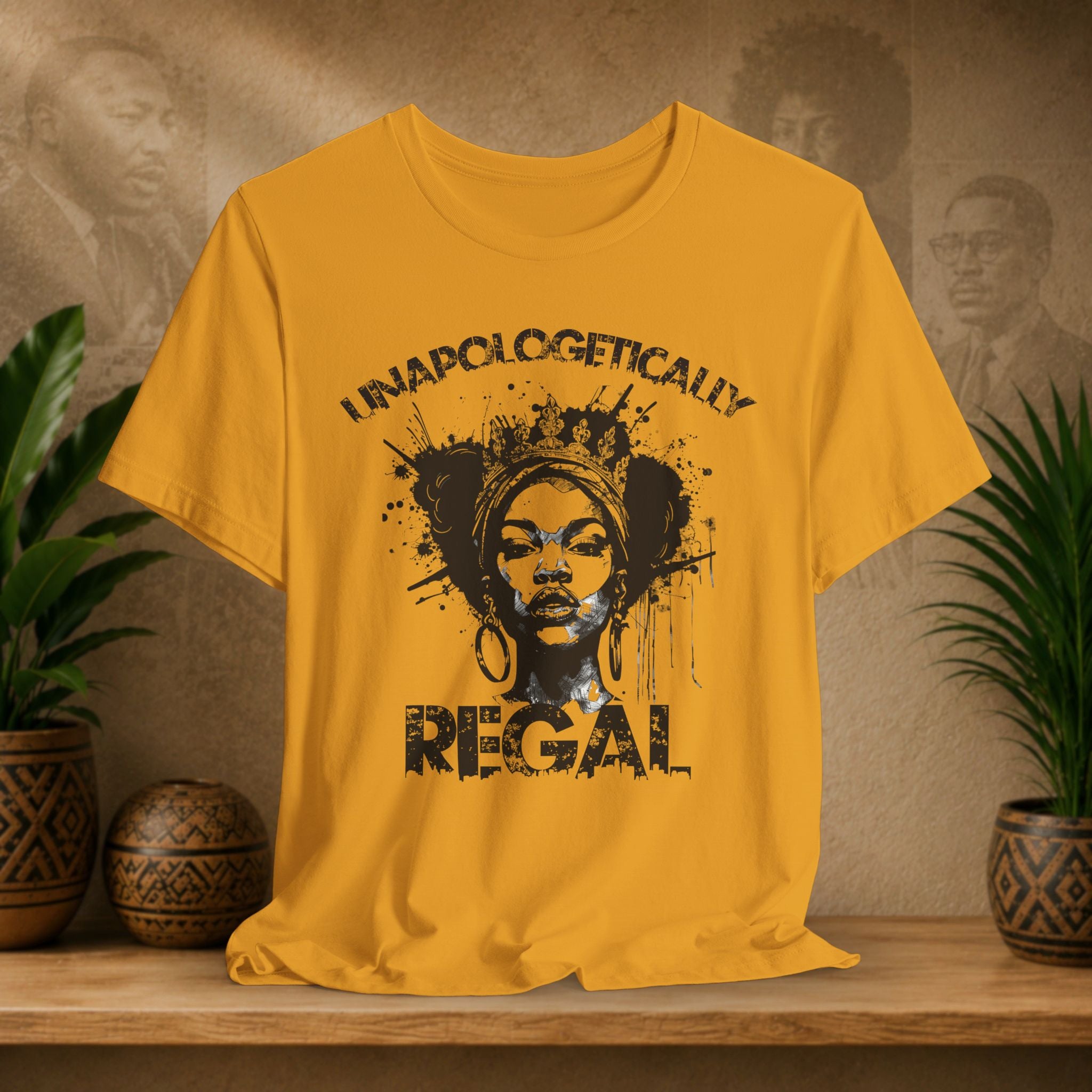 A mustard yellow short-sleeve graphic T-shirt displayed on a wooden surface, featuring a black distressed print of a woman's face with the words 'UNAPOLOGETICALLY REGAL.' The shirt is styled in a warm indoor setting with potted plants and a textured wall in the background.