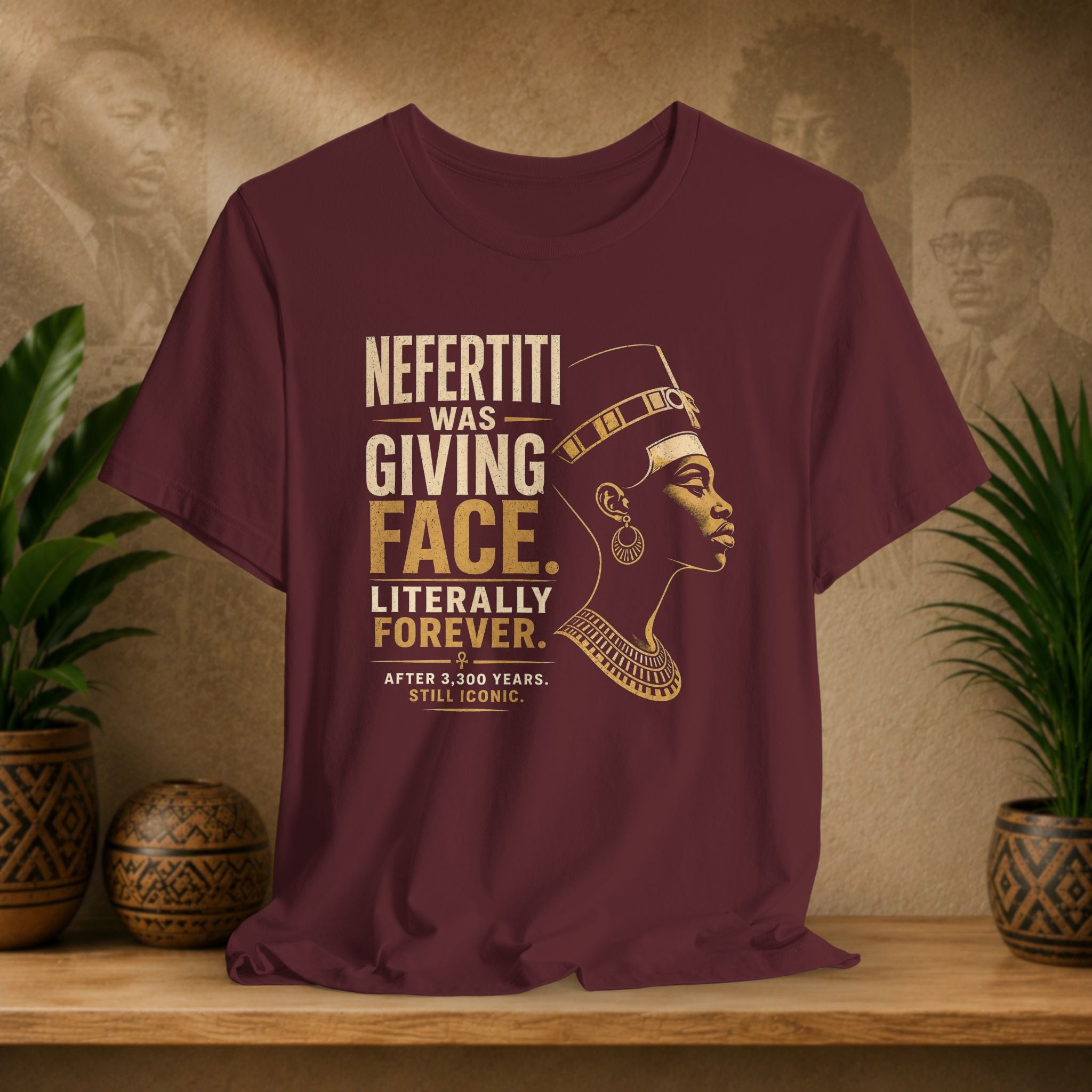 Maroon t-shirt with Nefertiti graphic and text on a wooden surface with decorative plants and background.