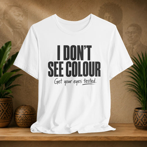 White t-shirt with text 'I DON'T SEE COLOUR Get your eyes tested.' on a wooden surface with plants and a textured wall background.