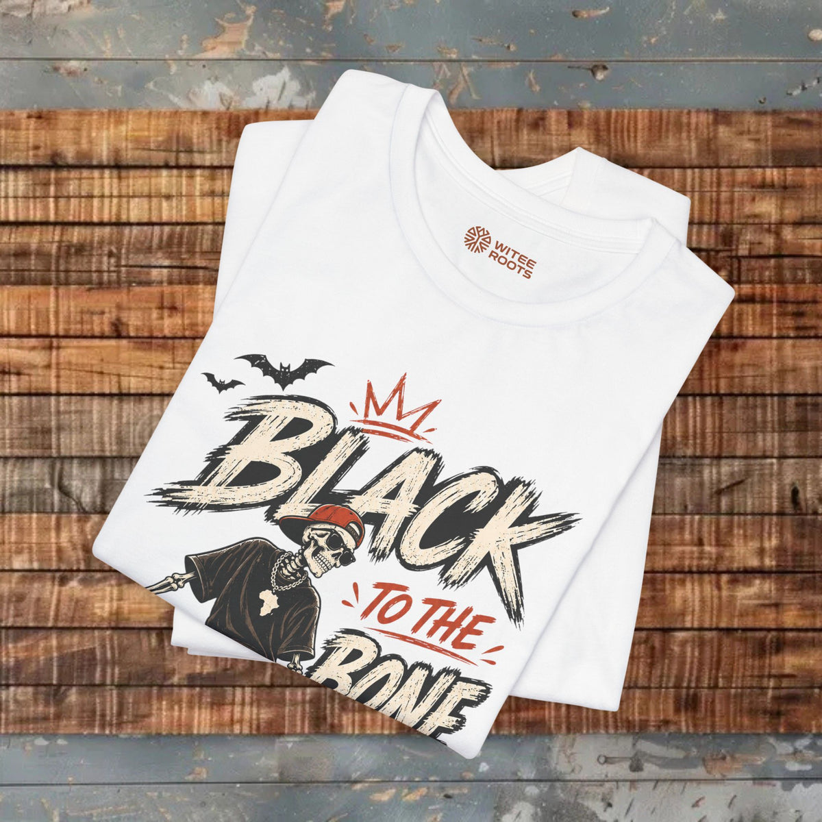 A folded white graphic T-shirt displayed on a rustic wooden plank surface. The shirt features a black, white, and red graphic with the text 'BLACK TO THE RONE' and a skeleton wearing a red Santa hat, with two black bats above.