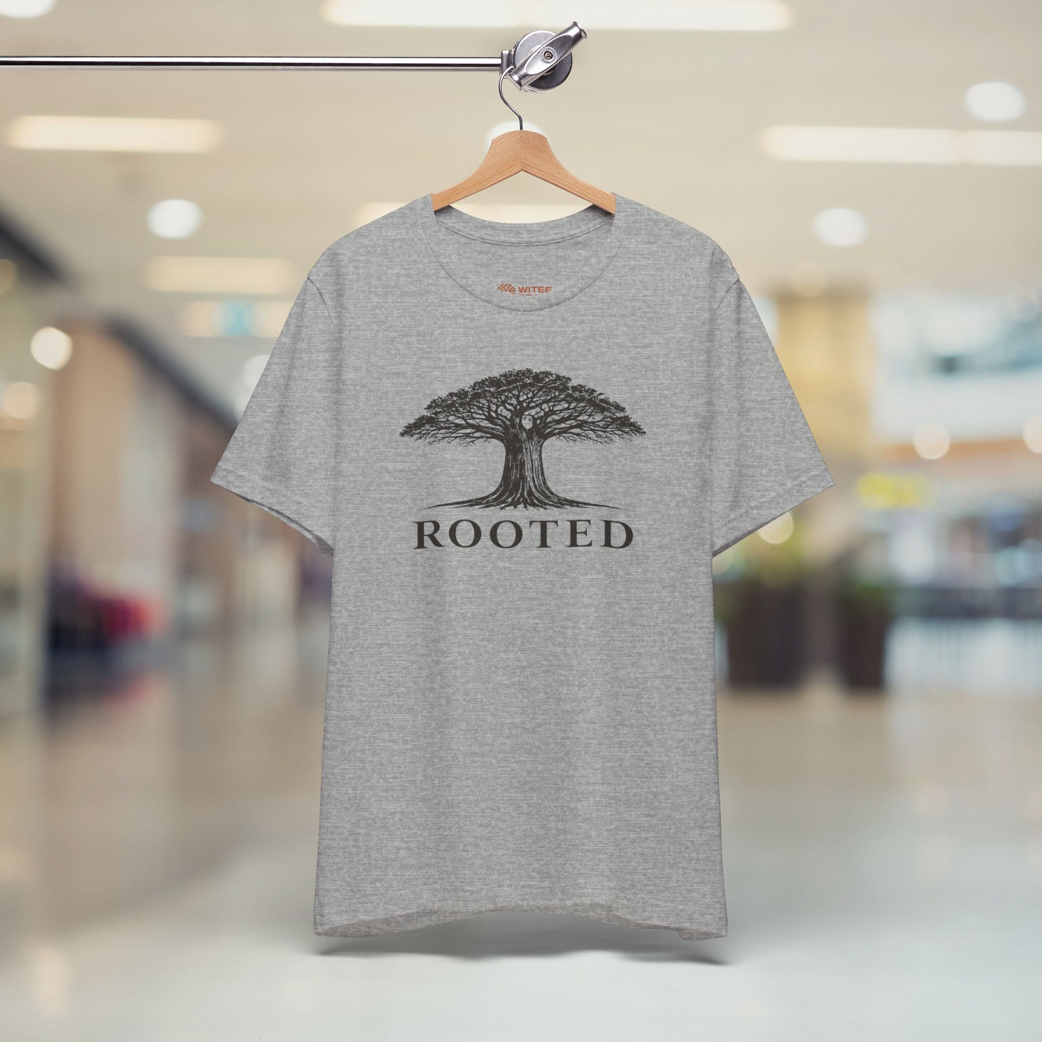 Gray t-shirt with a tree graphic and 'ROOTED' text on a hanger in a blurred indoor setting