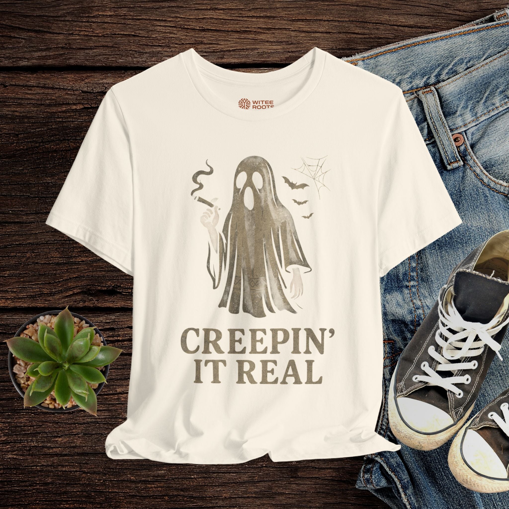 Beige t-shirt with ghost graphic and 'Creepin' It Real' text on a wooden surface with jeans and sneakers.