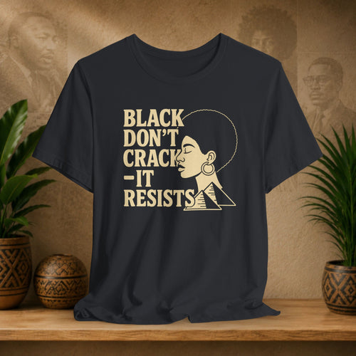 Black t-shirt with 'Black Don't Crack, It Resists' text and graphic on a wooden surface with plants and decorative items.