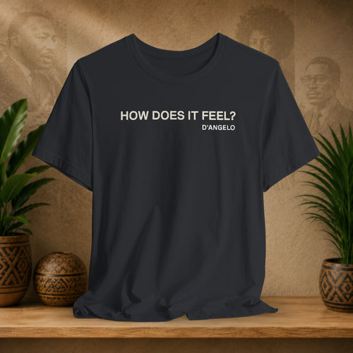 Black t-shirt with 'HOW DOES IT FEEL? D'ANGELO' text on a wooden shelf with plants and decorative items.
