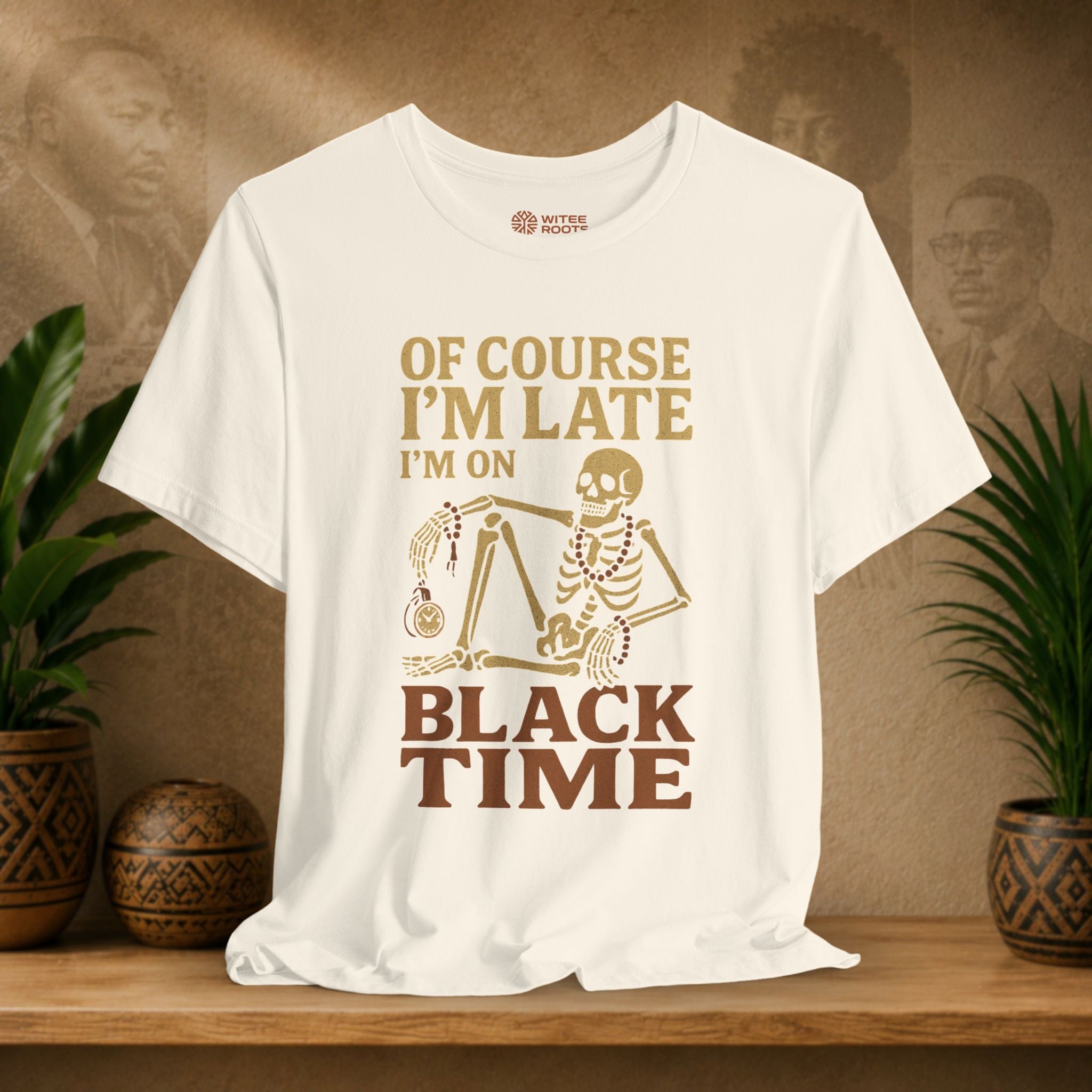A beige graphic t-shirt displayed on a wooden shelf, featuring a brown and gold design with the text 'OF COURSE I'M LATE I'M ON BLACK TIME' and a skeleton holding a clock. The background is a warm brown wall with subtle portraits, flanked by potted green plants.