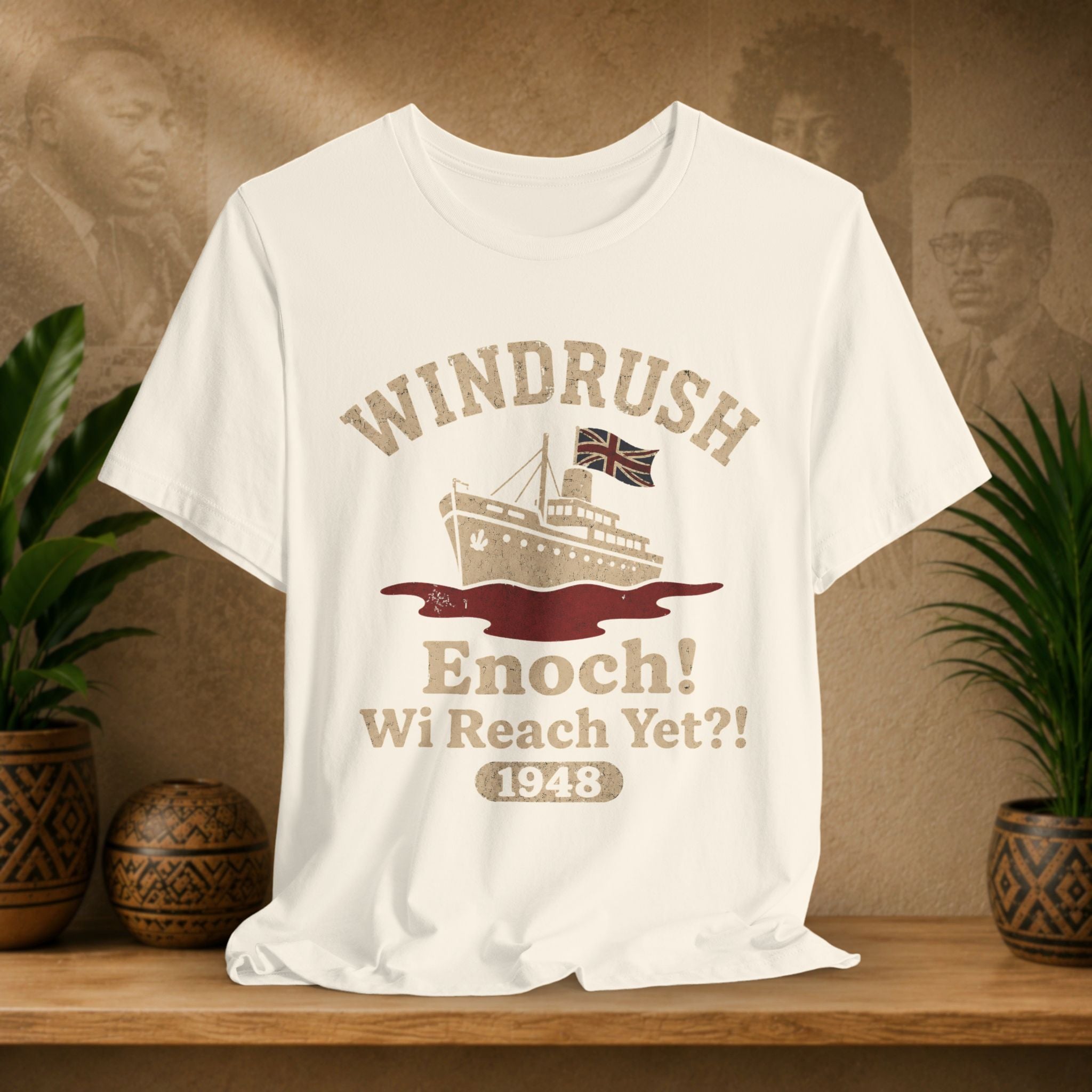 Beige t-shirt with 'Windrush Enoch! Wi Reach Yet?? 1948' design on a wooden surface with plants and a vintage-style background.