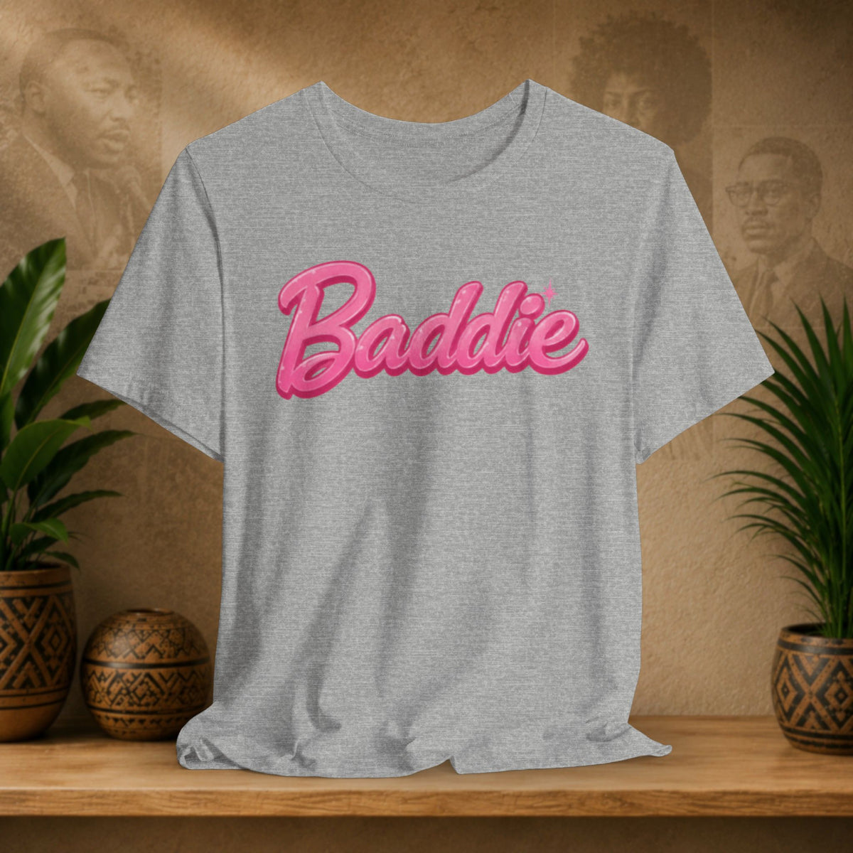 A heather gray short-sleeve t-shirt with the word 'Baddie' printed across the chest in a pink 3D-effect script font, styled on a wooden shelf with potted plants and a textured background.