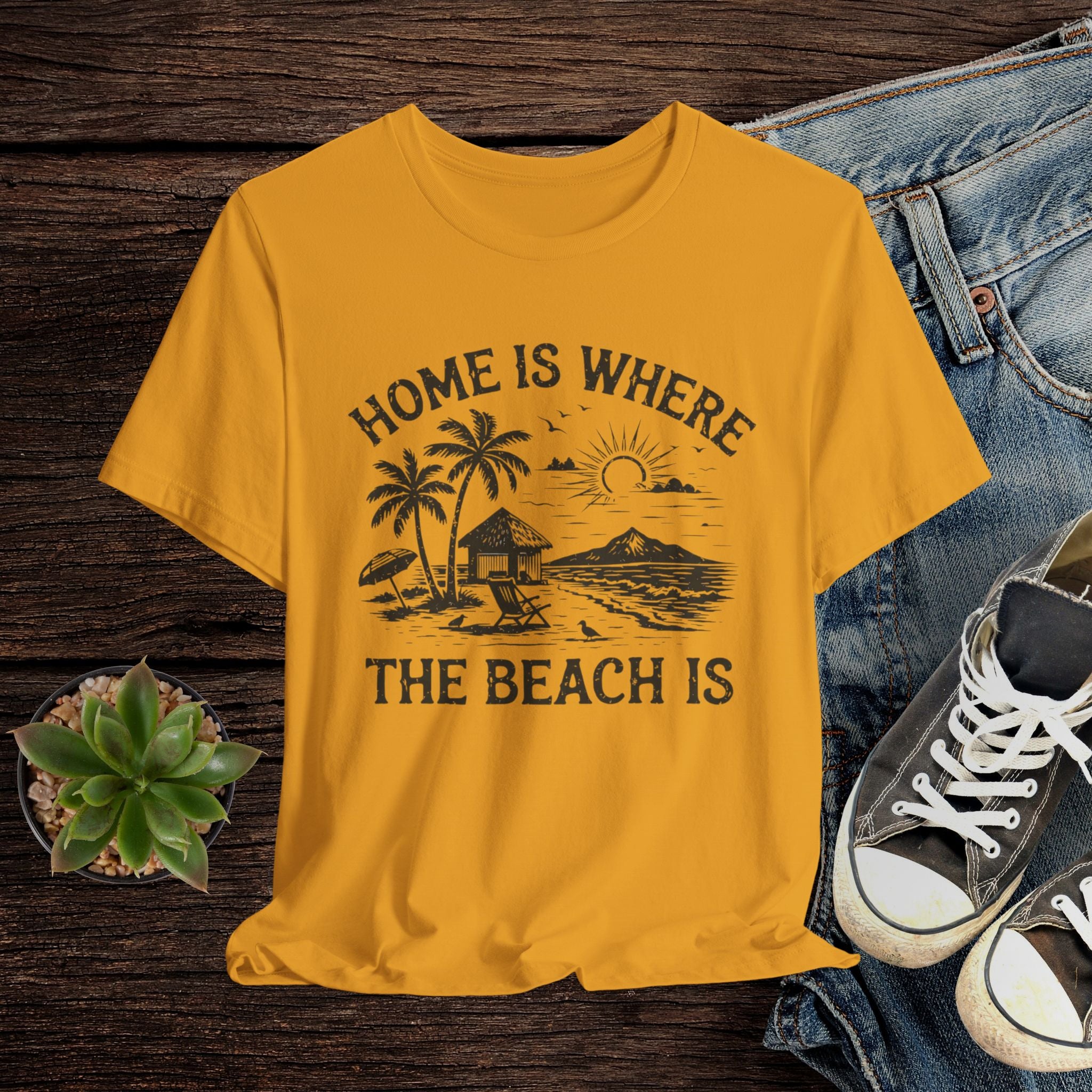 Mustard t-shirt with beach graphic and text on a wooden surface with jeans and sneakers.