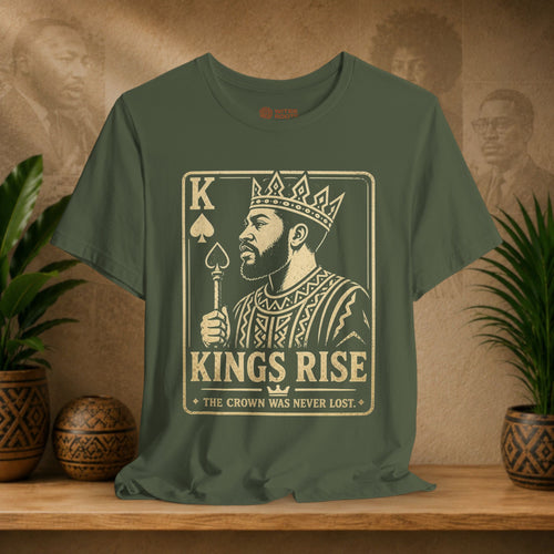 Green t-shirt with a graphic design of a king holding a sword and the text 'KINGS RISE' on a wooden surface with plants and a decorative wall.