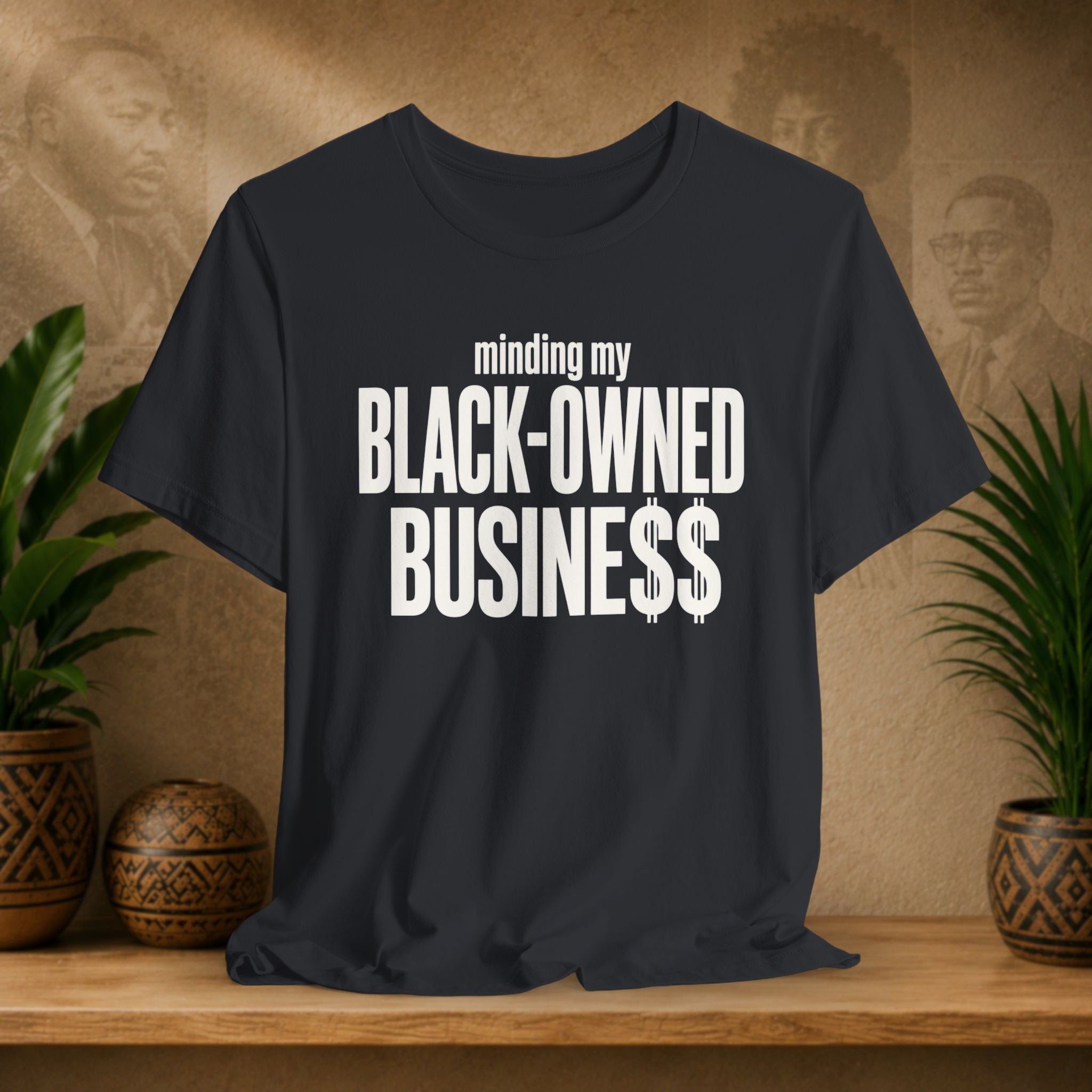 Black t-shirt with bold white text reading 'minding my BLACK-OWNED BUSINESS$$' displayed on a wooden shelf with decorative plants and textured background.