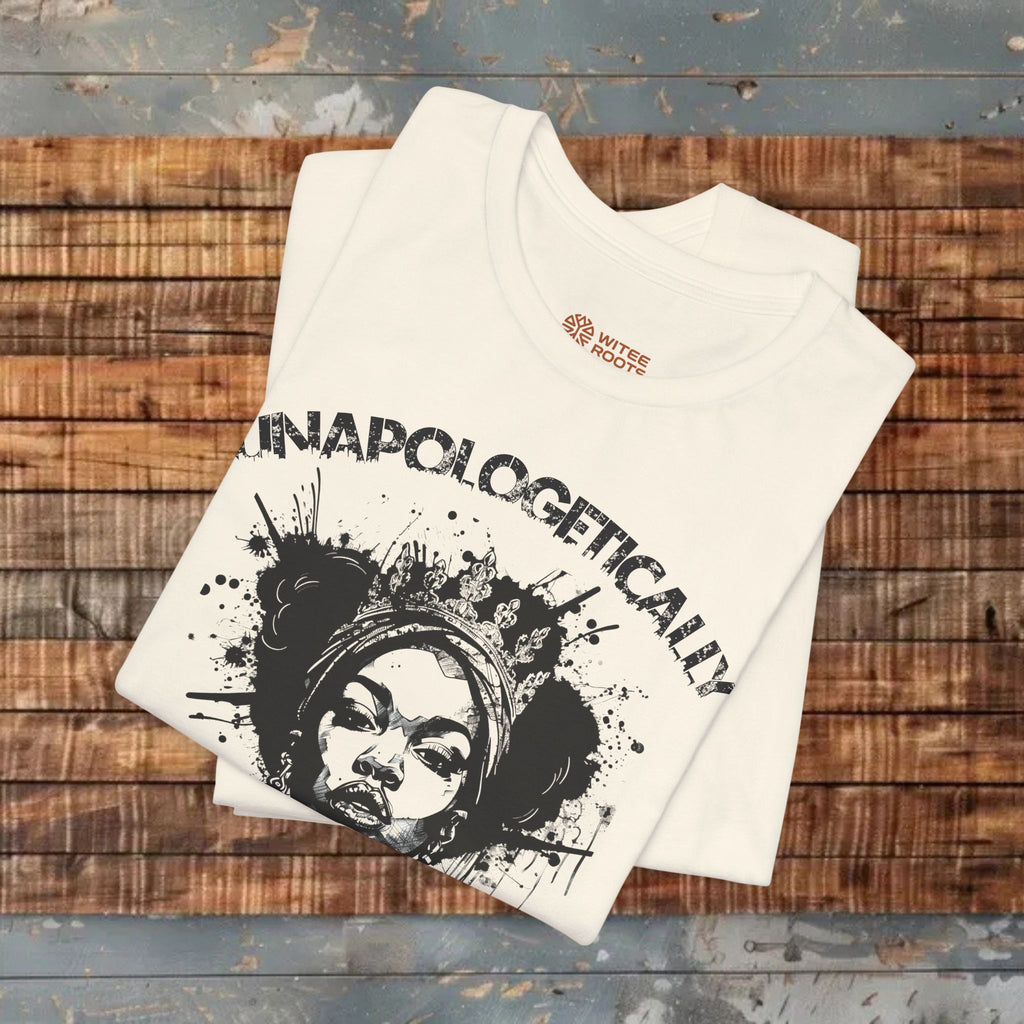 A folded off-white graphic T-shirt with a black illustrated face and the word 'UNAPOLOGETICALLY' printed across the front, displayed on a rustic wooden plank surface. A small 'WITNES STUDIOS' logo is visible inside the collar.